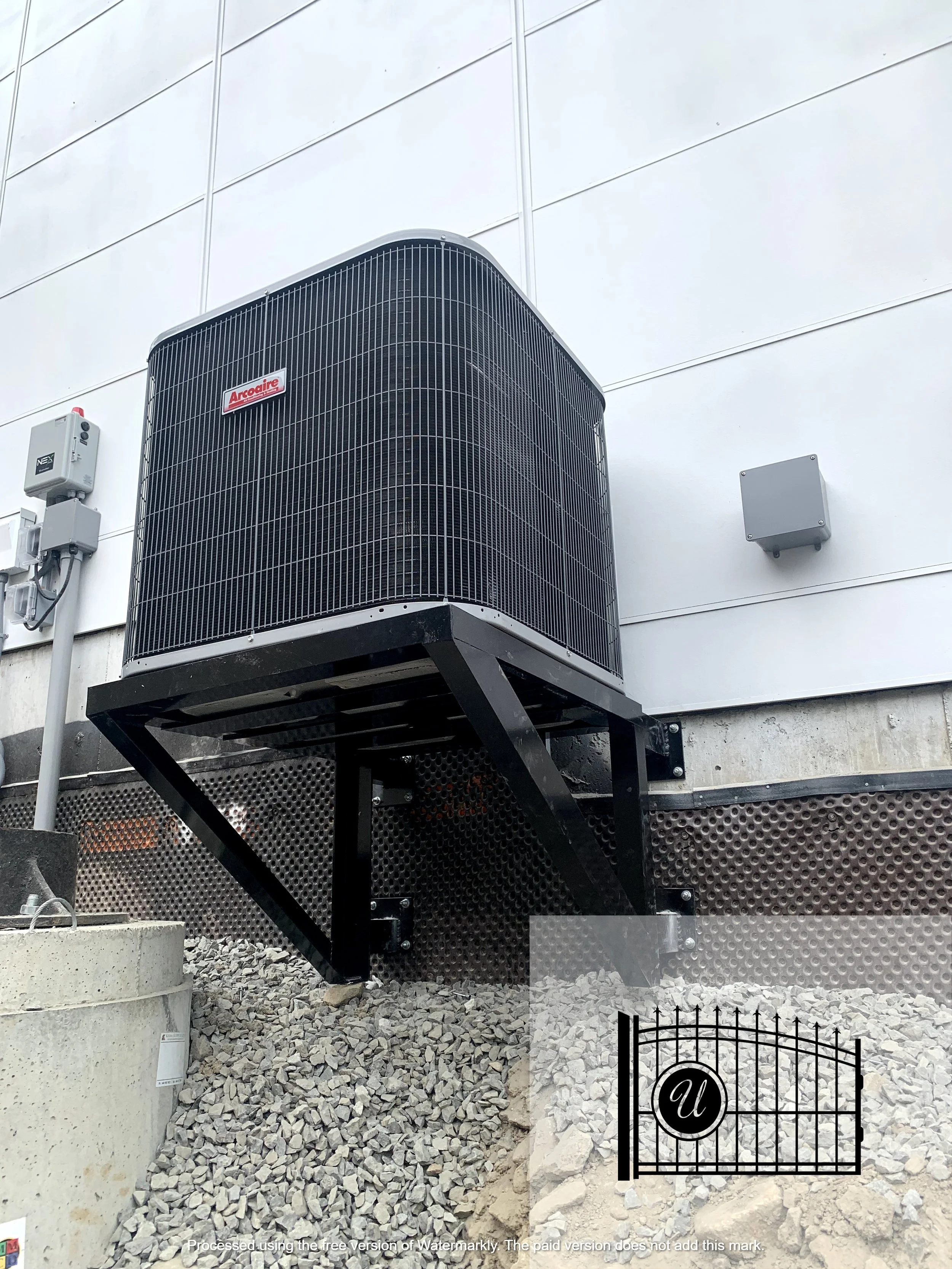 Outdoor air conditioning unit mounted on a wall, with electrical components nearby, on a building exterior with white siding and gravel ground.