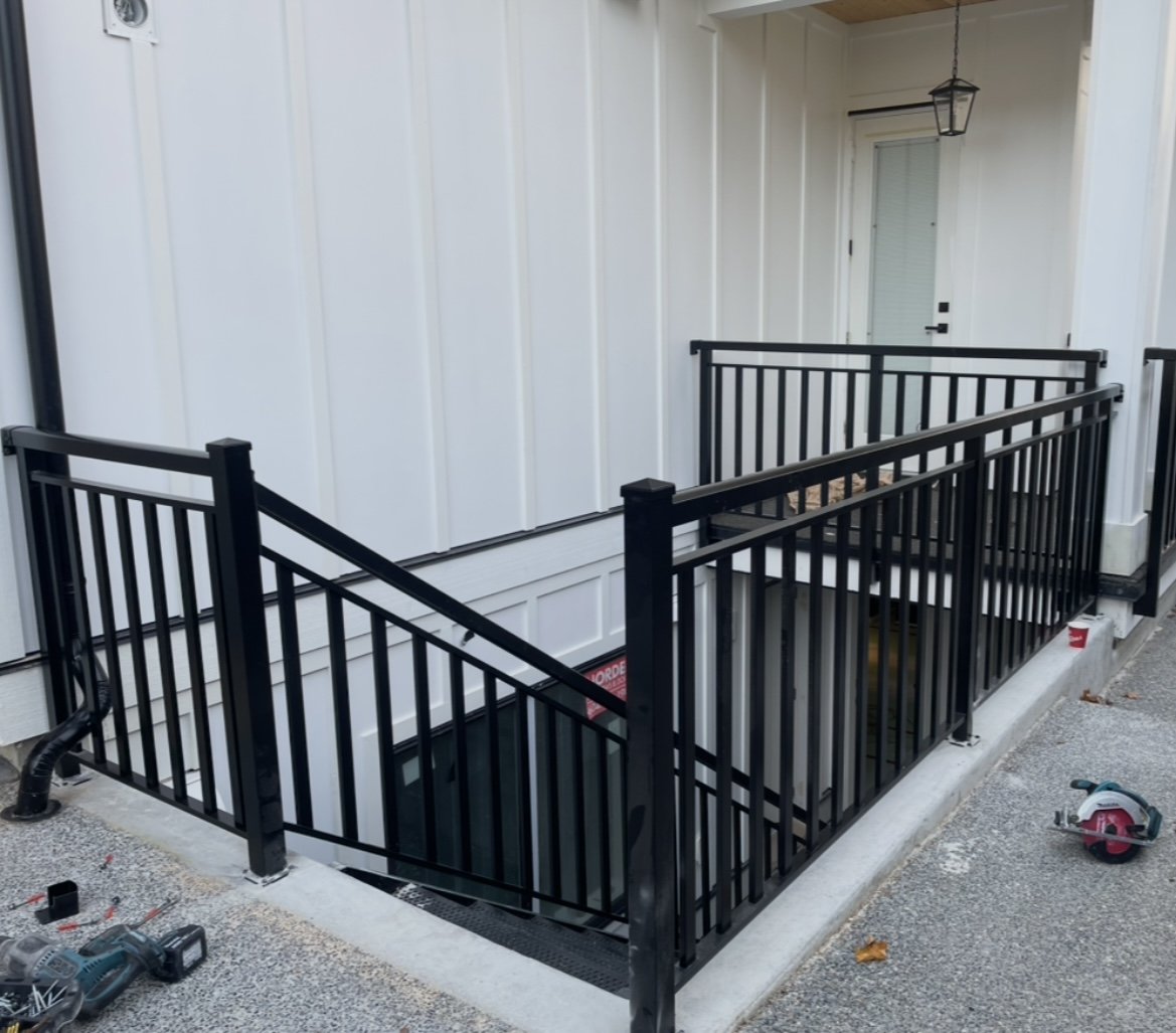 A black metal safety railing surrounds an outdoor basement staircase entrance to a house with white siding. Construction tools and materials are nearby.