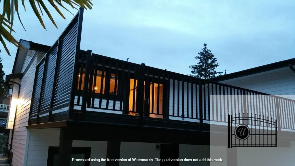A house with a black balcony railing, illuminated interior, and trees in the background during dusk.