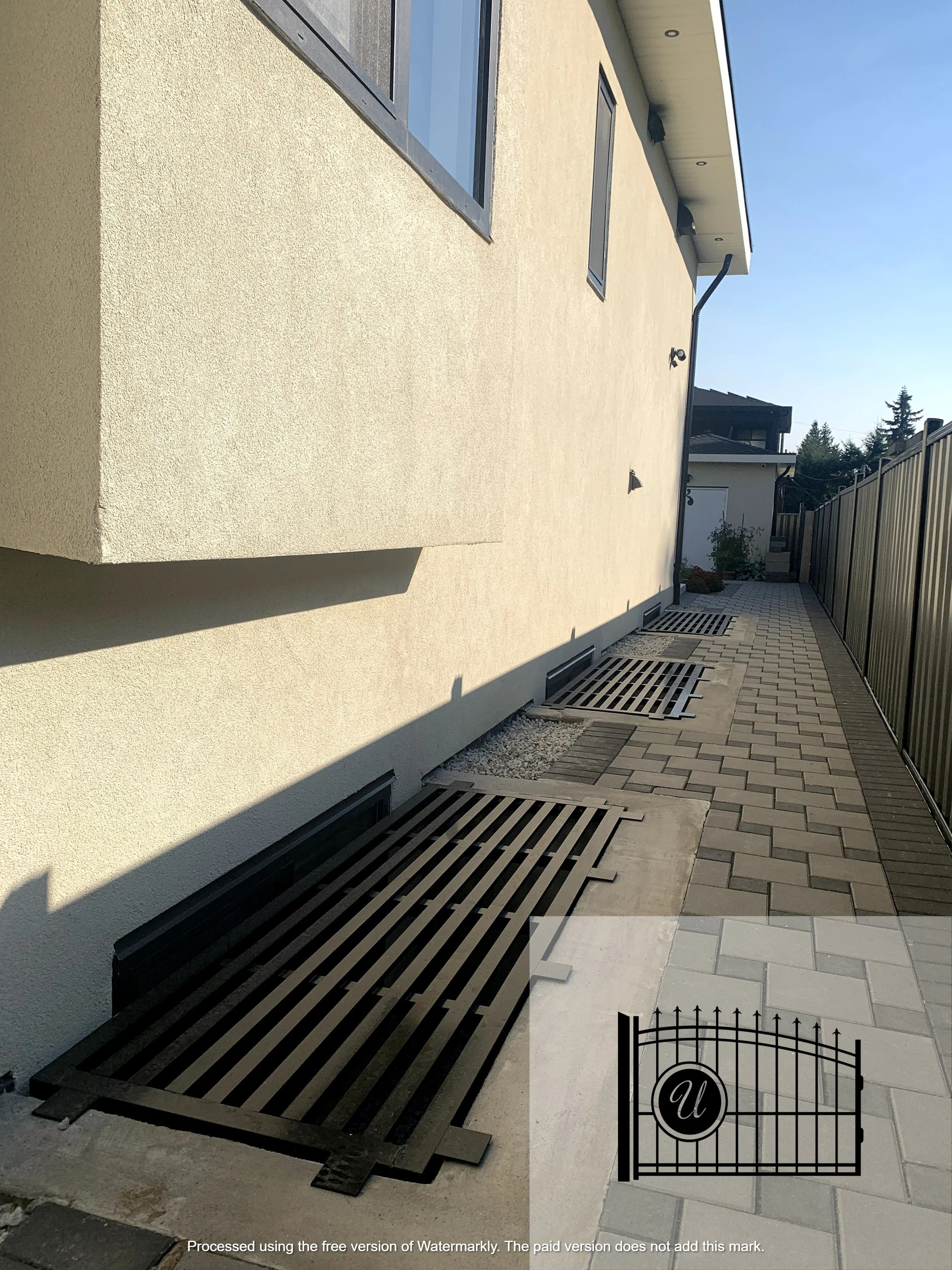 A beige stucco exterior wall with a black metal gate in the foreground and a paved pathway along the side of the house. There are windows and roof vents visible on the house, with a blue sky above.
