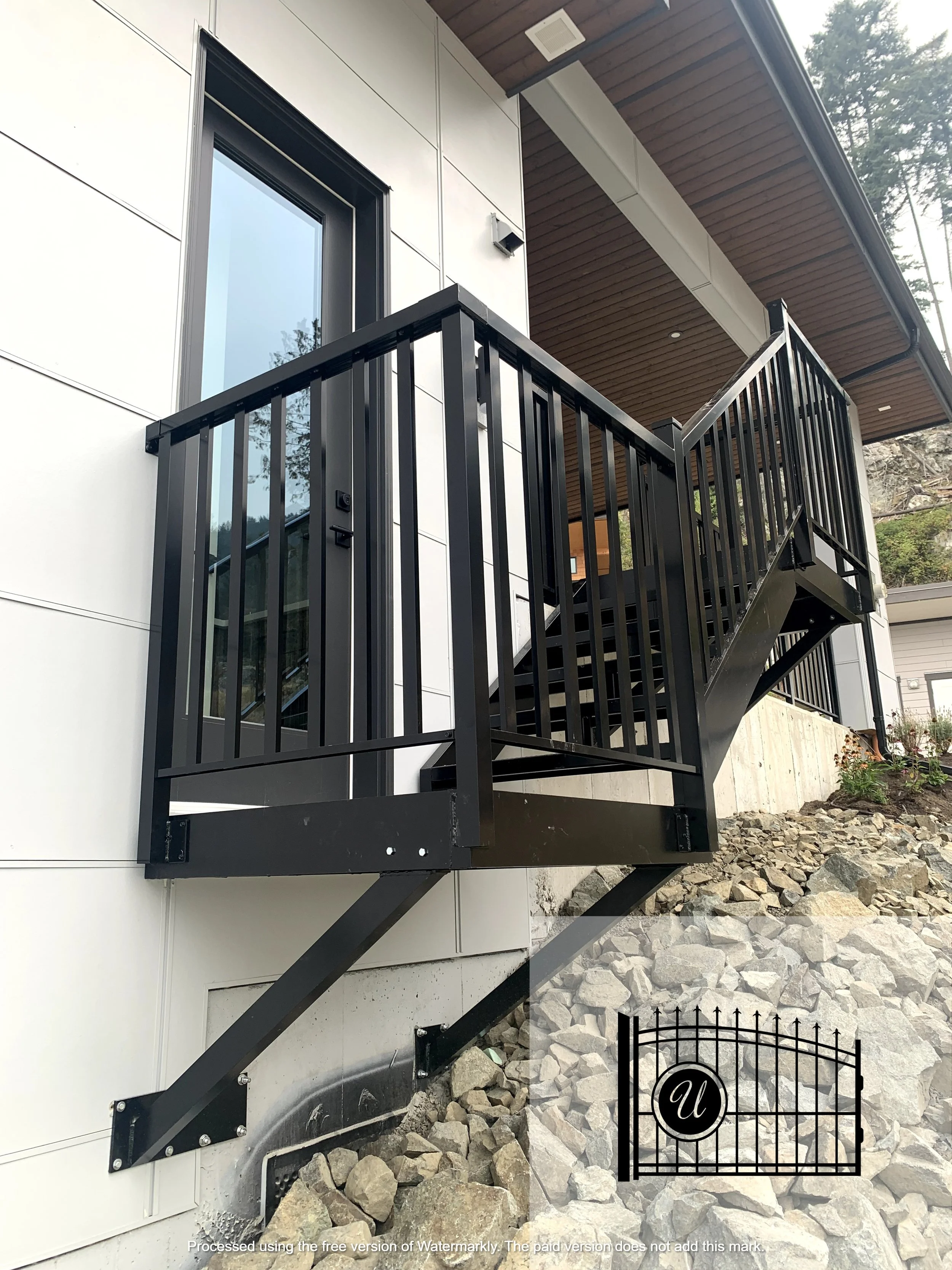 Exterior view of a modern house with a black metal staircase leading to a balcony with a black railing, white siding, and a wooden ceiling, surrounded by rocks and trees.