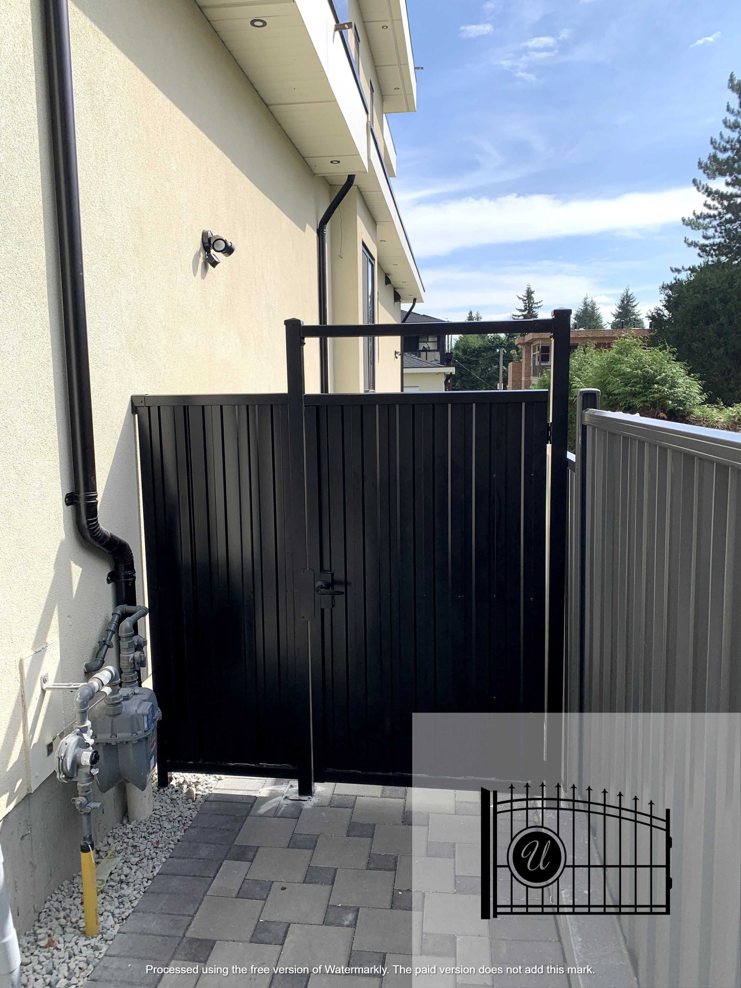 A black metal gate with a decorative letter 'U' at the bottom right, in front of a beige house wall with an outdoor surveillance camera and gas meter, and a partly cloudy sky in the background.