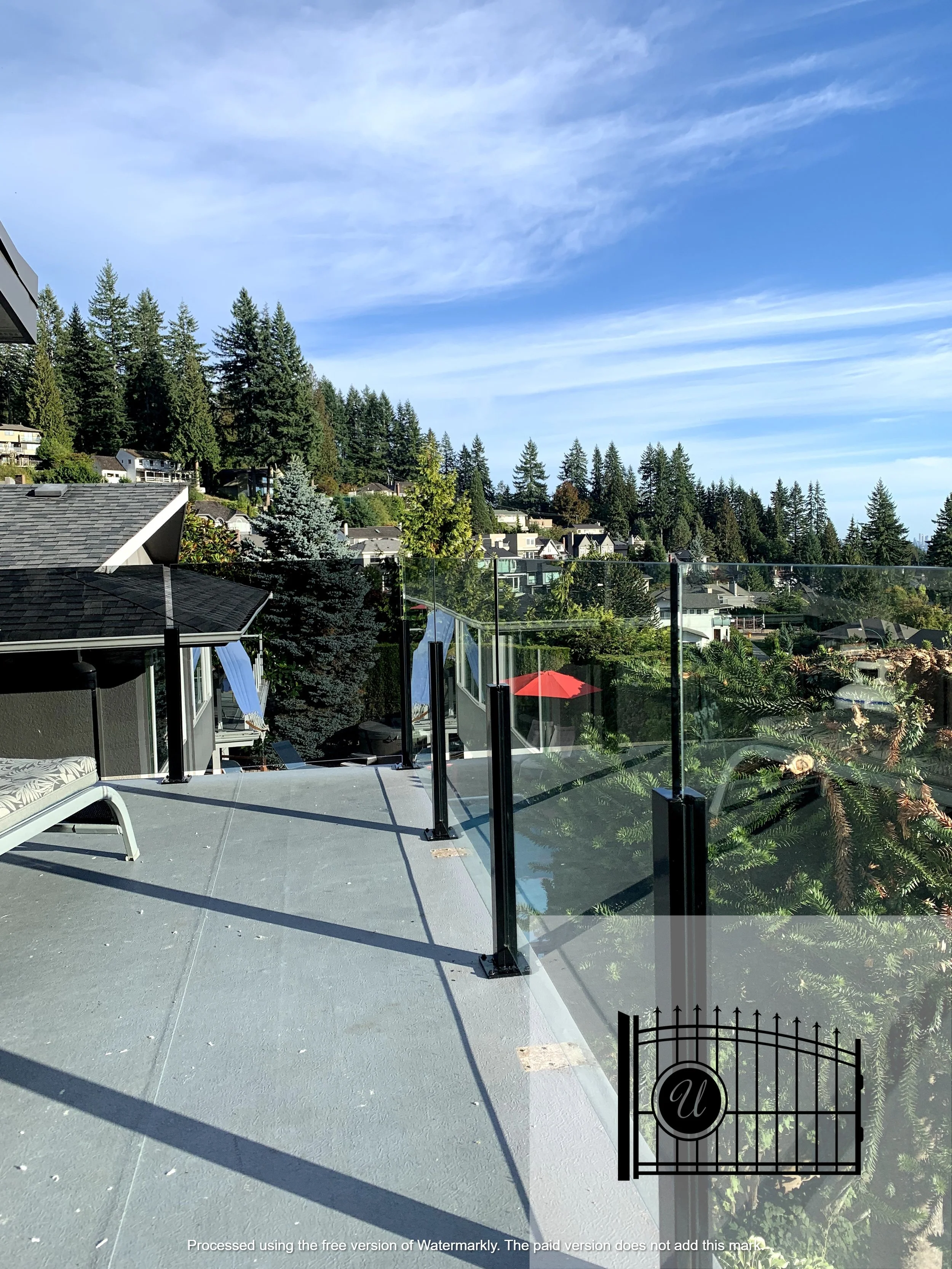 A suburban backyard patio with a lounge chair, a glass railing, and a view of houses and tall trees under a blue sky with scattered clouds.