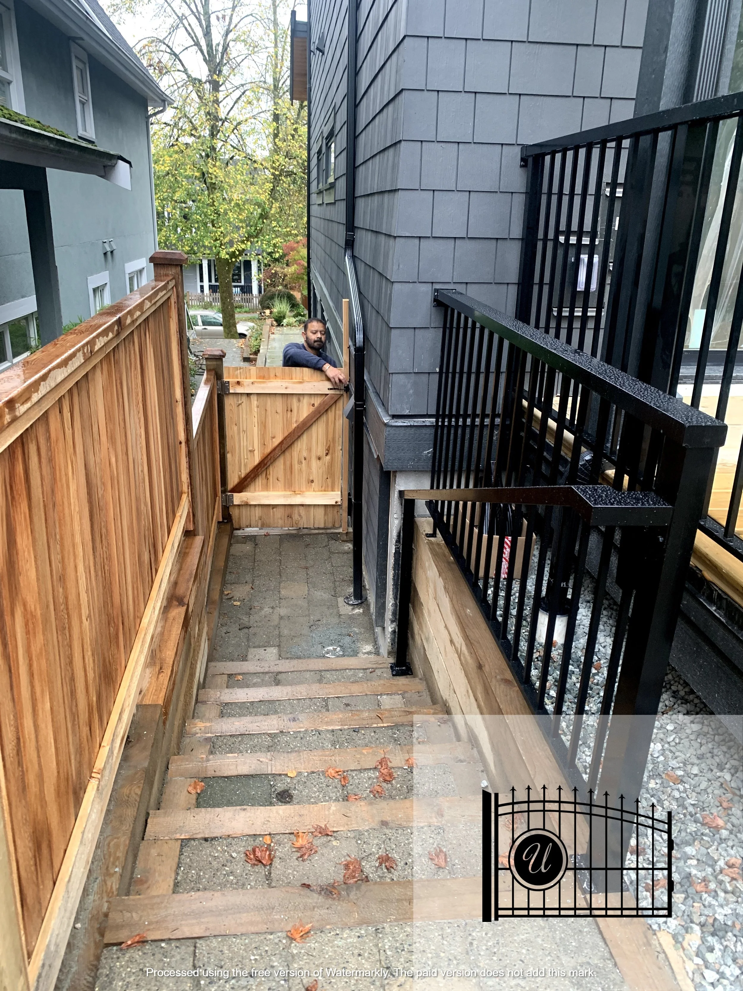 A man standing behind a wooden gate at the top of a newly built staircase, with the staircase missing some steps. The scene is outdoors between two houses, one with blue siding and the other with gray shingles, with trees in the background.