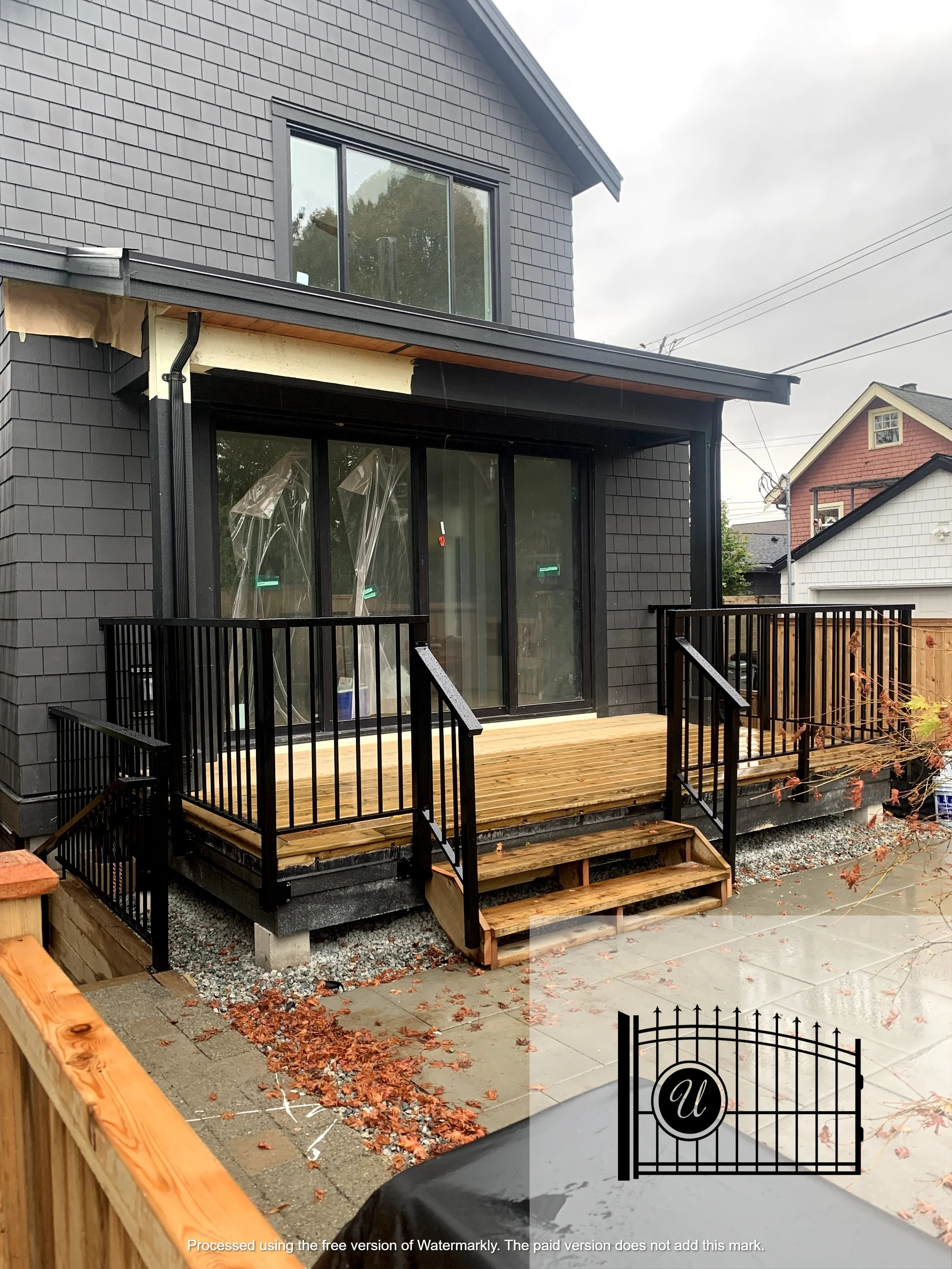 A residential house under construction with a small wooden deck and black metal railing, concrete steps, a black metal gate, and fallen autumn leaves on the ground. The house is gray with shingle siding, large windows, and a newer extension with slid