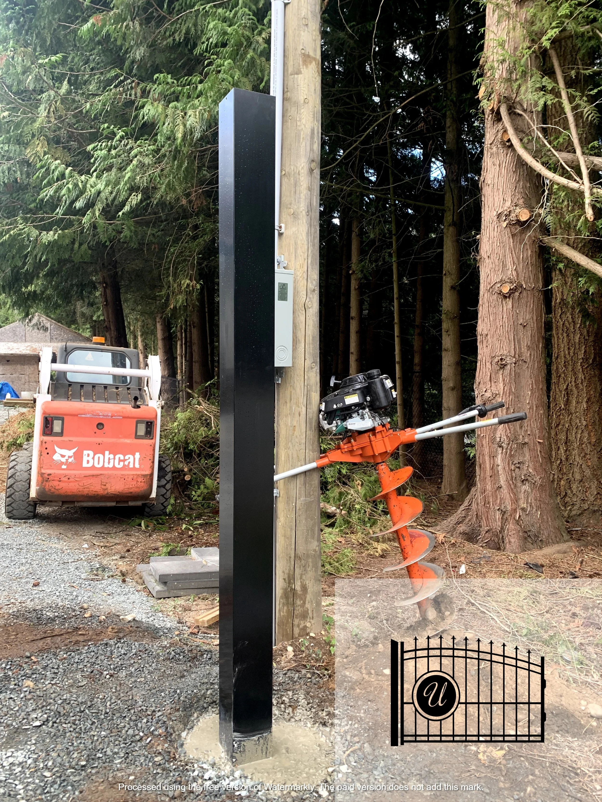 A black electric vehicle charging station mounted on a wooden utility pole next to a large tree, with an orange auger tool leaning against the tree and a Bobcat loader in the background.