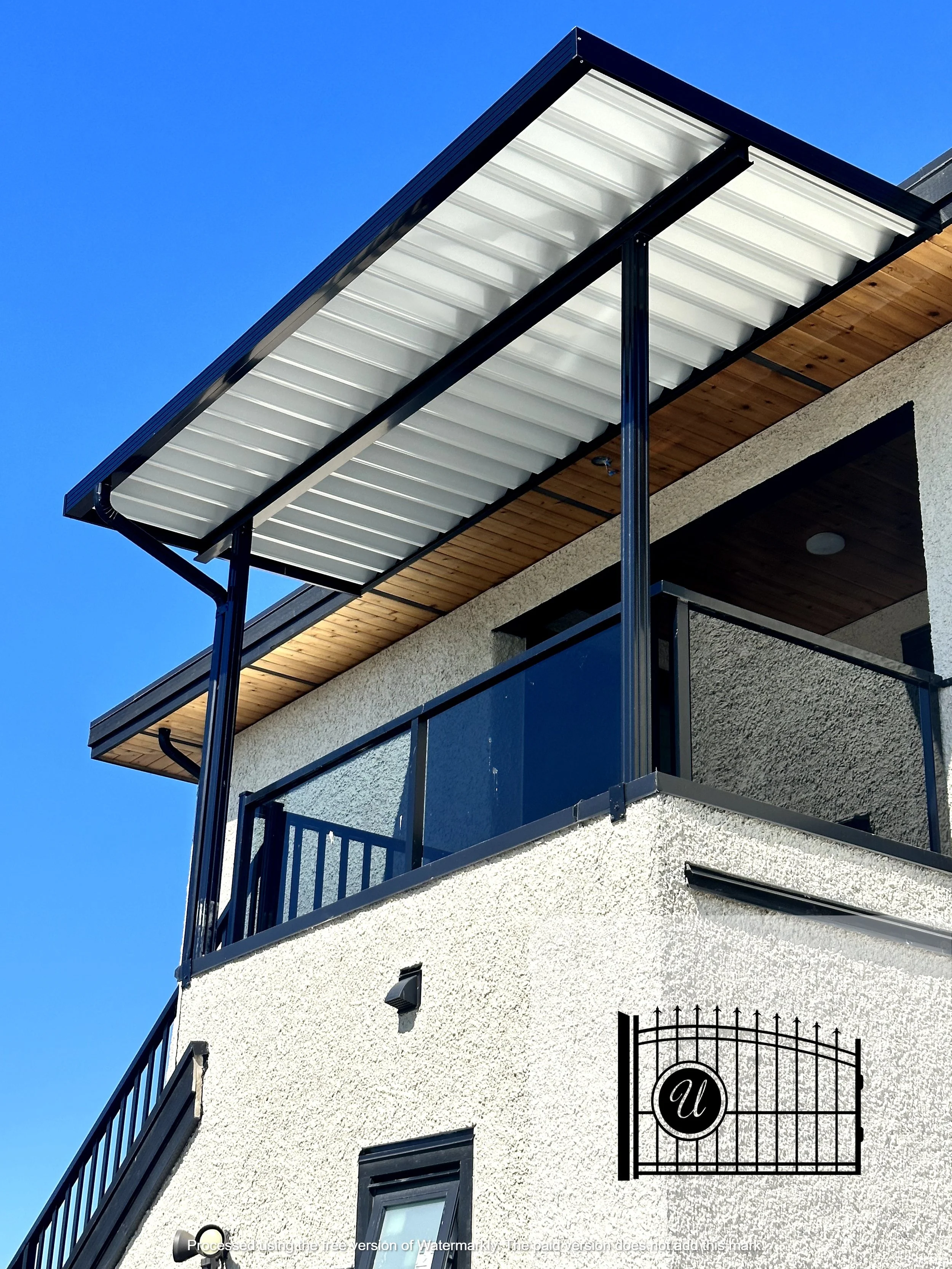 Exterior view of a modern house with a balcony, black metal railing, and a solar panel on the roof, against a clear blue sky.