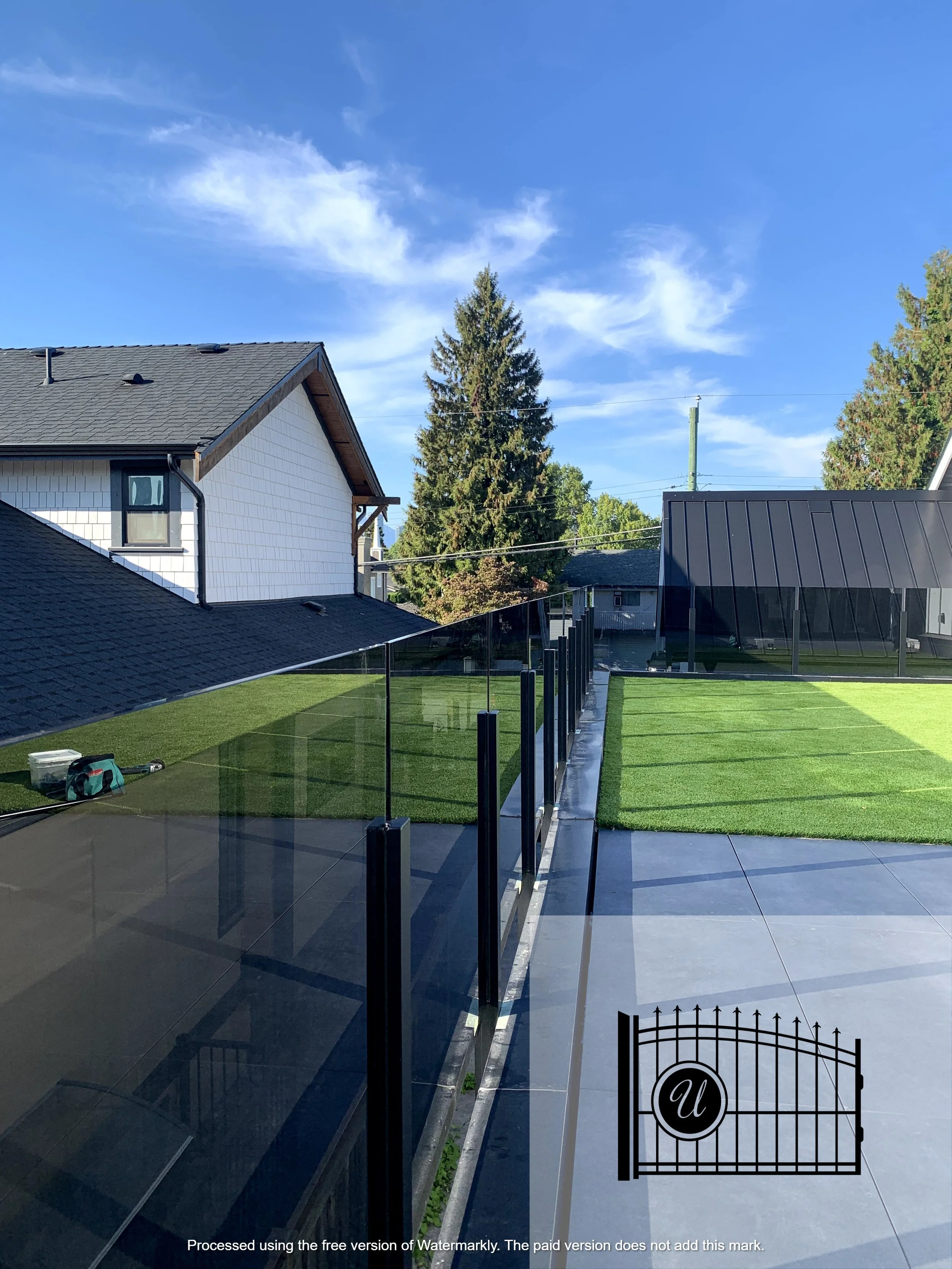 A backyard with artificial grass, a black metal fence, and a house with white siding and a black roof. There is a large evergreen tree and other trees in the background, along with a partly cloudy blue sky.