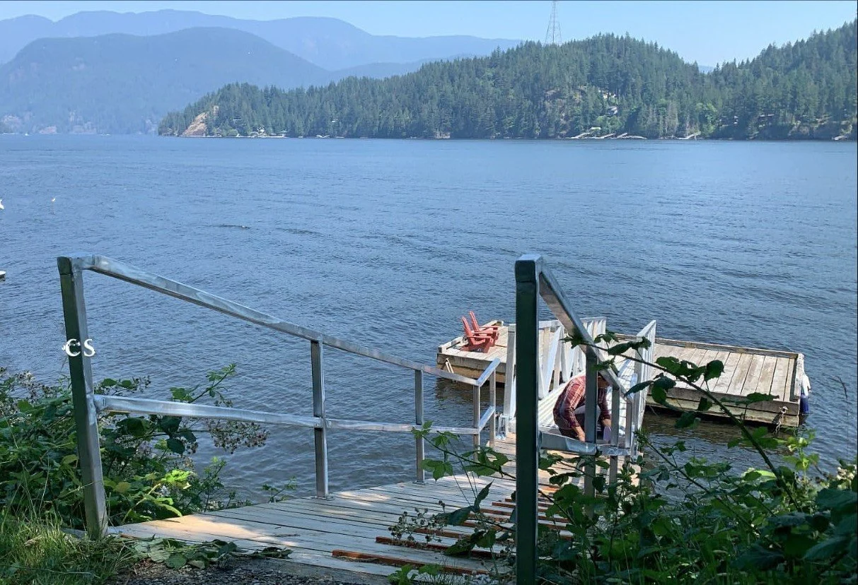 A lakeside scene with a metal staircase leading down to a small wooden dock. Two red chairs and some white chairs are on the dock, overlooking the calm water of a large lake. Forested hills and mountains are visible in the background under a clear bl