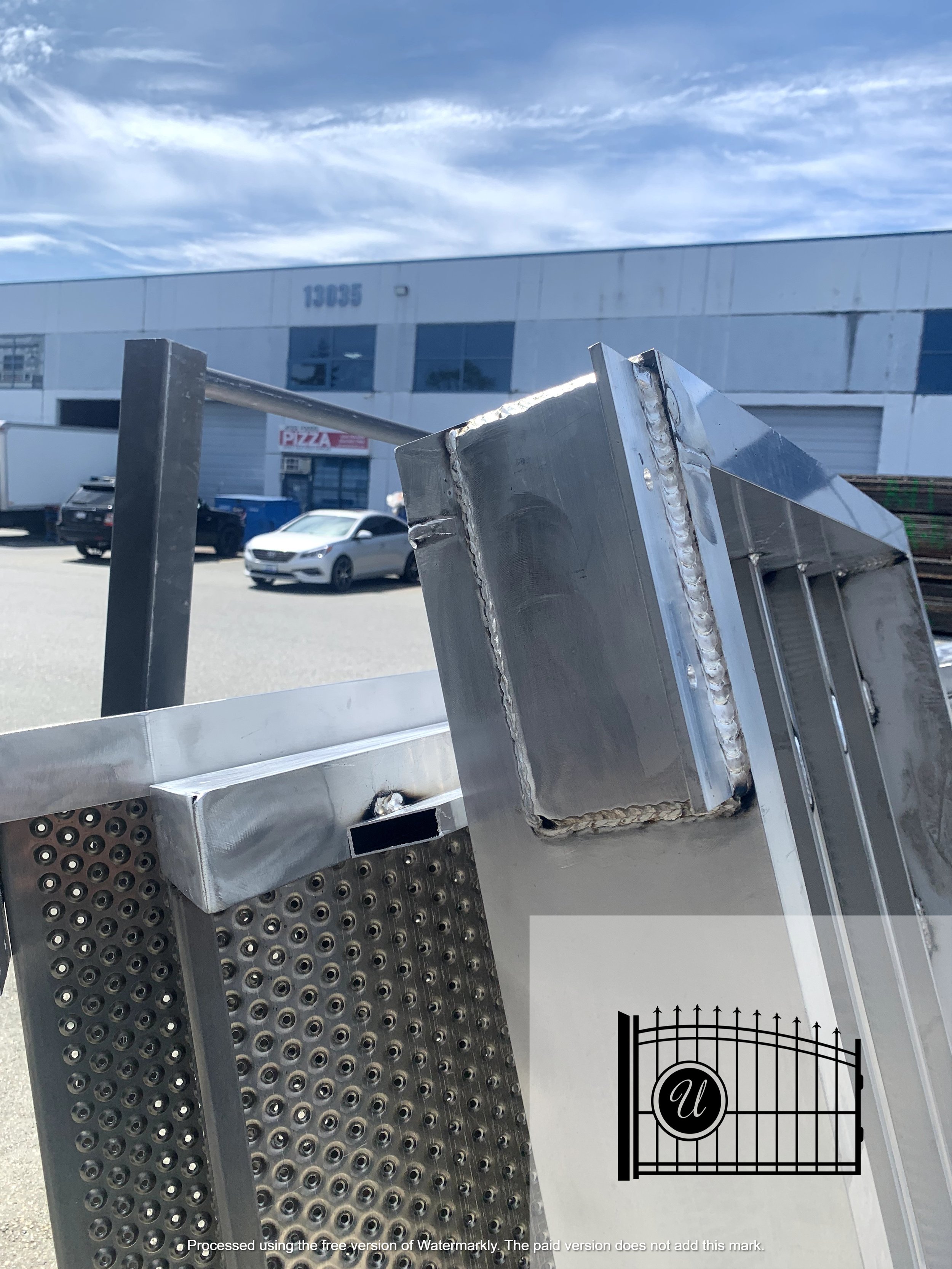 Close-up of a metal construction or industrial equipment with welded joints and perforated panels, outdoors in front of commercial-industrial buildings and parked cars under a cloudy sky.