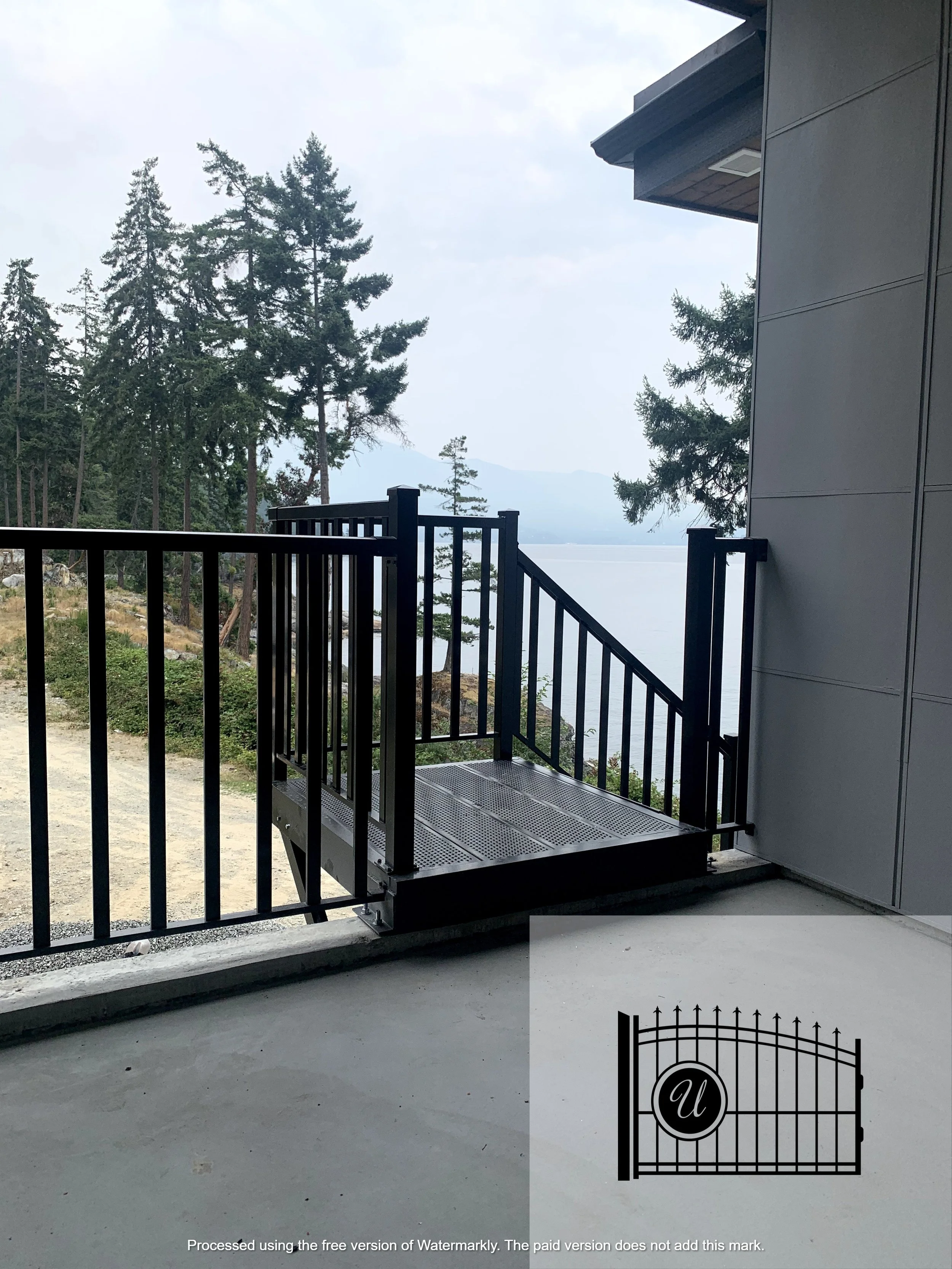 Photo of a balcony with black railings overlooking a forested area with tall trees and mountains in the distance, under a cloudy sky.