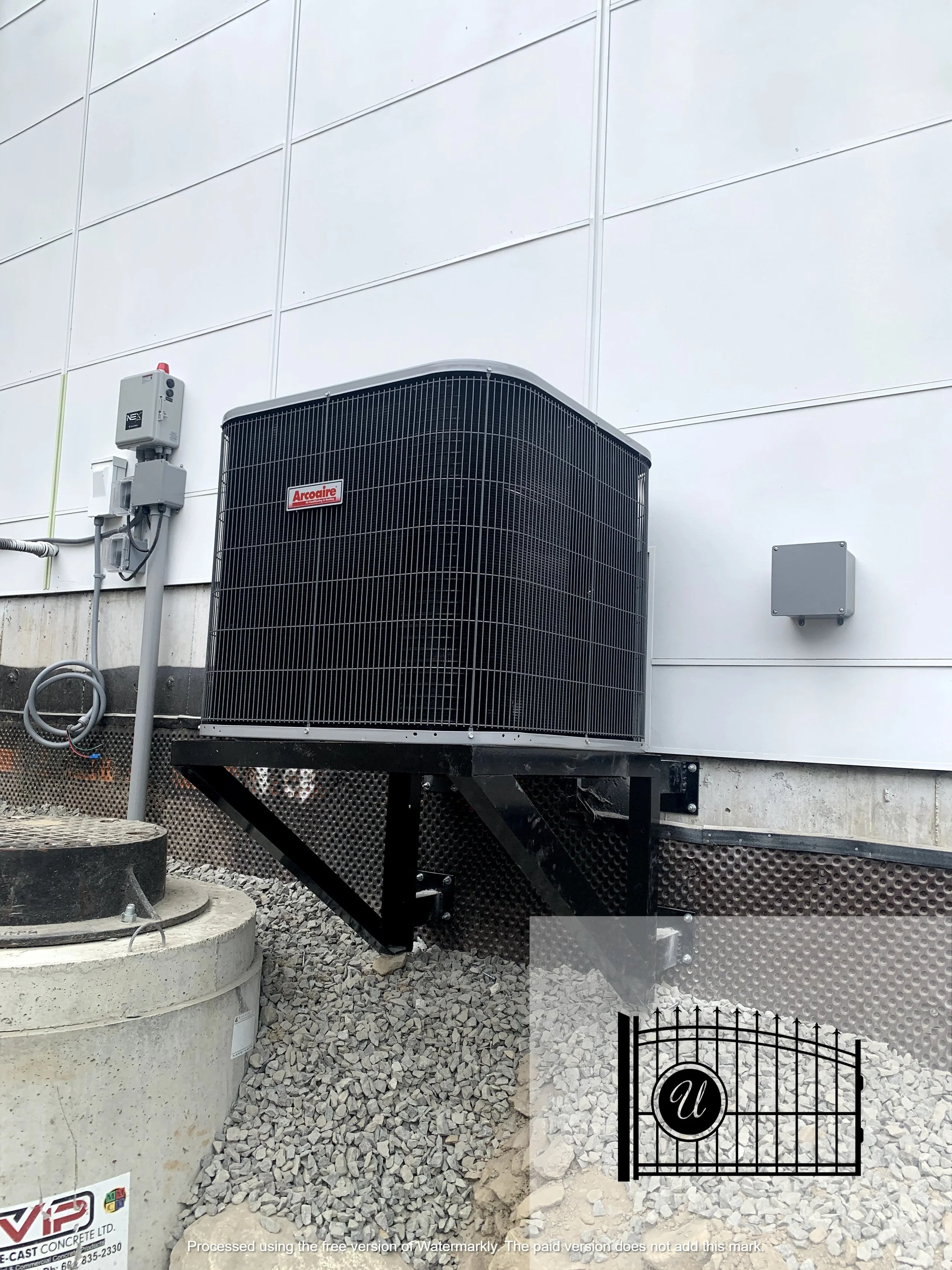 An air conditioning unit mounted on a metal stand outside a building with white panel walls, electrical wiring, and a small fenced area with gravel and a concrete base.
