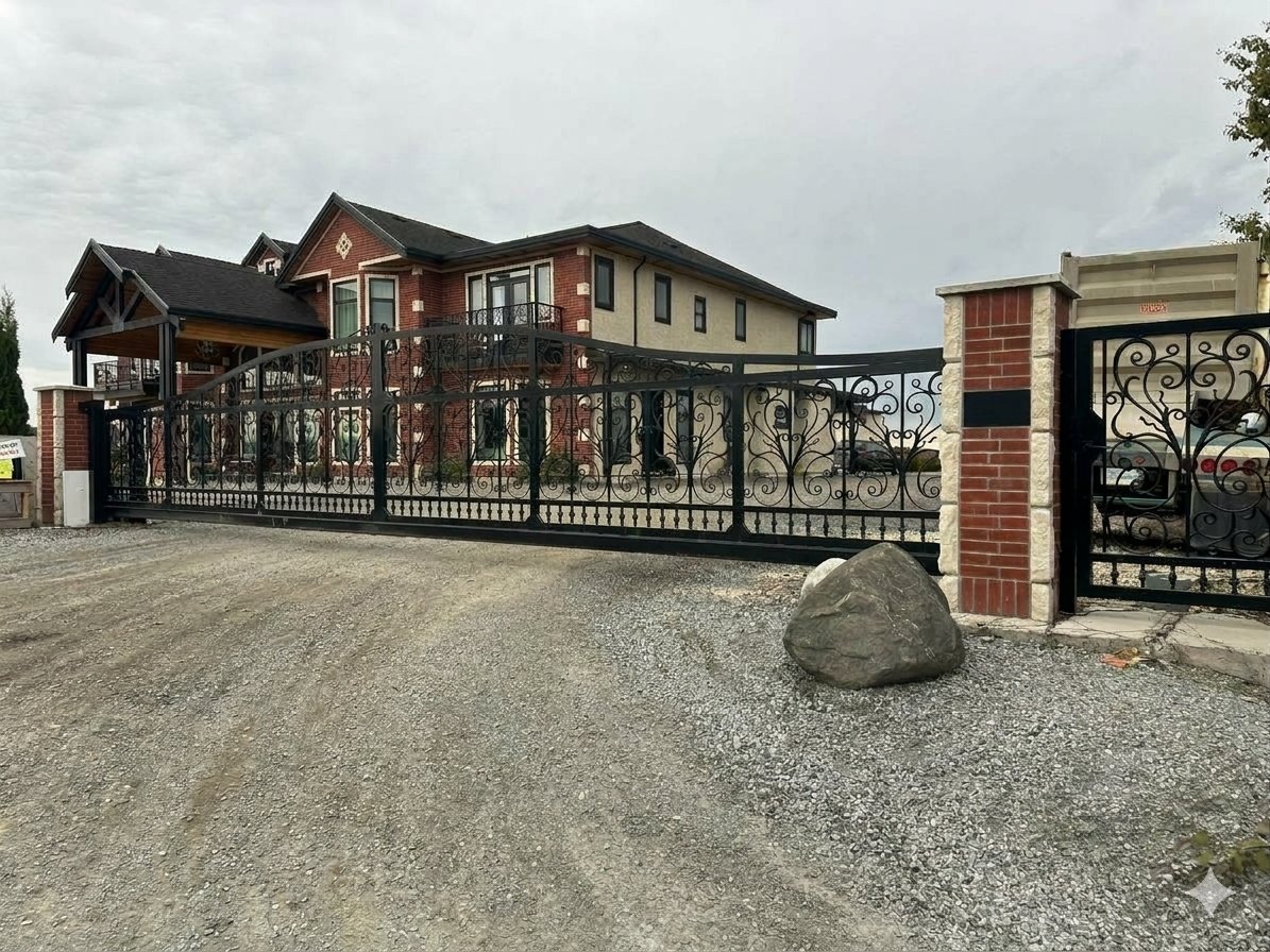A large house behind an ornate black wrought iron gate with brick pillars, a gravel driveway, and parked cars, under an overcast sky.
