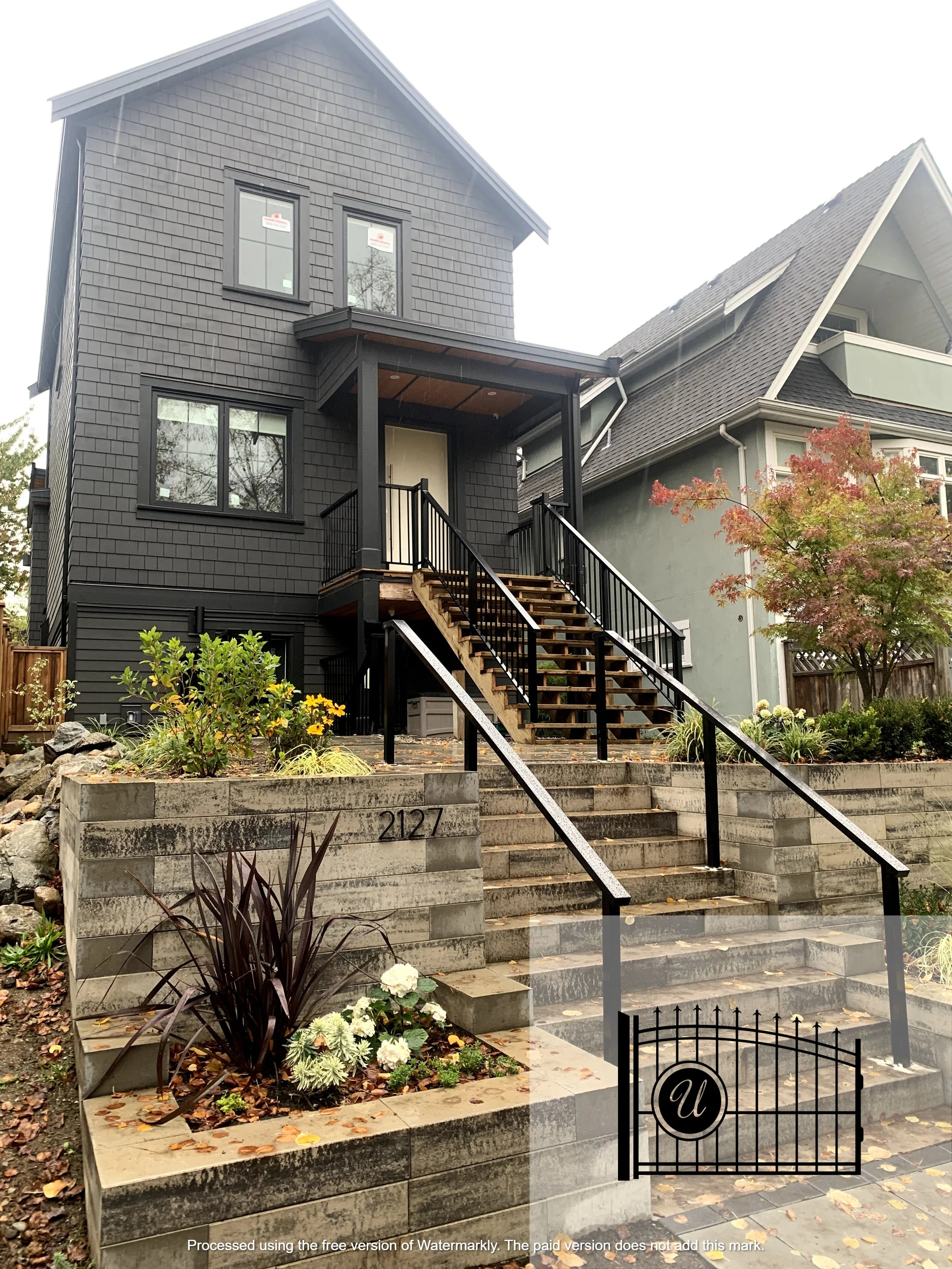 A modern black-painted house with multiple windows and a staircase leading to the front door, surrounded by a small garden with plants and flowers, and a black gate with a circular emblem.