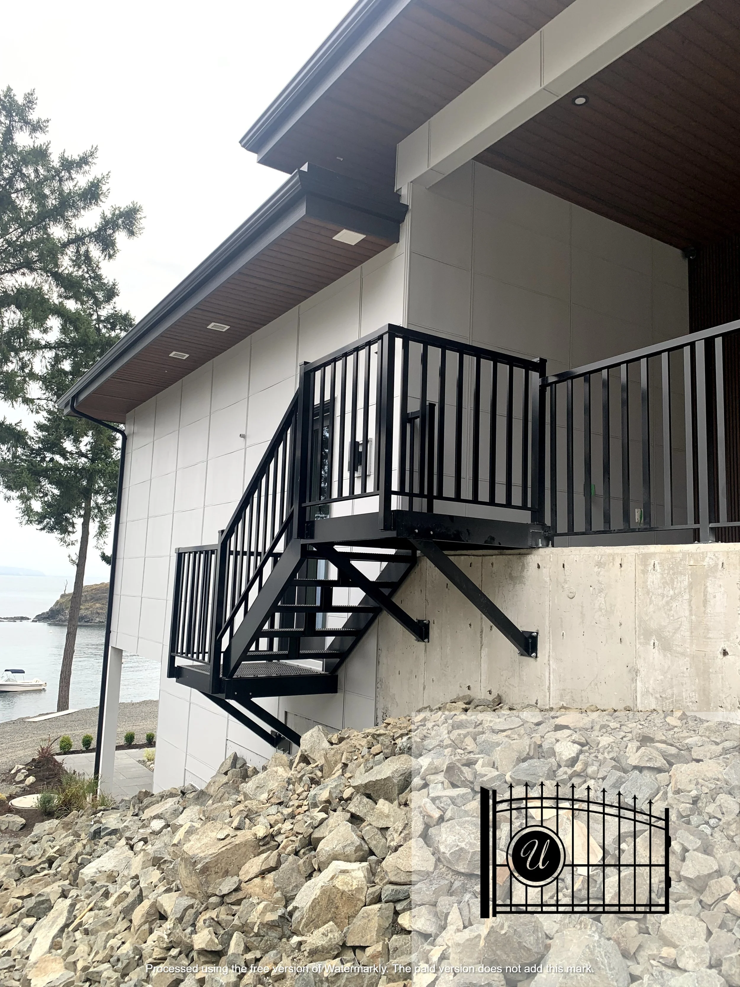 Modern house exterior with metal staircase, large rocks, and a view of water and trees in the background.