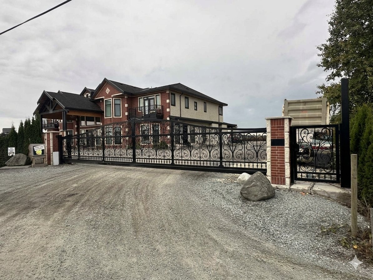 A large two-story house with a gated driveway, brick and beige exterior, and ornate black metal fencing. Gravel driveway with rocks and parked cars visible behind the gate.