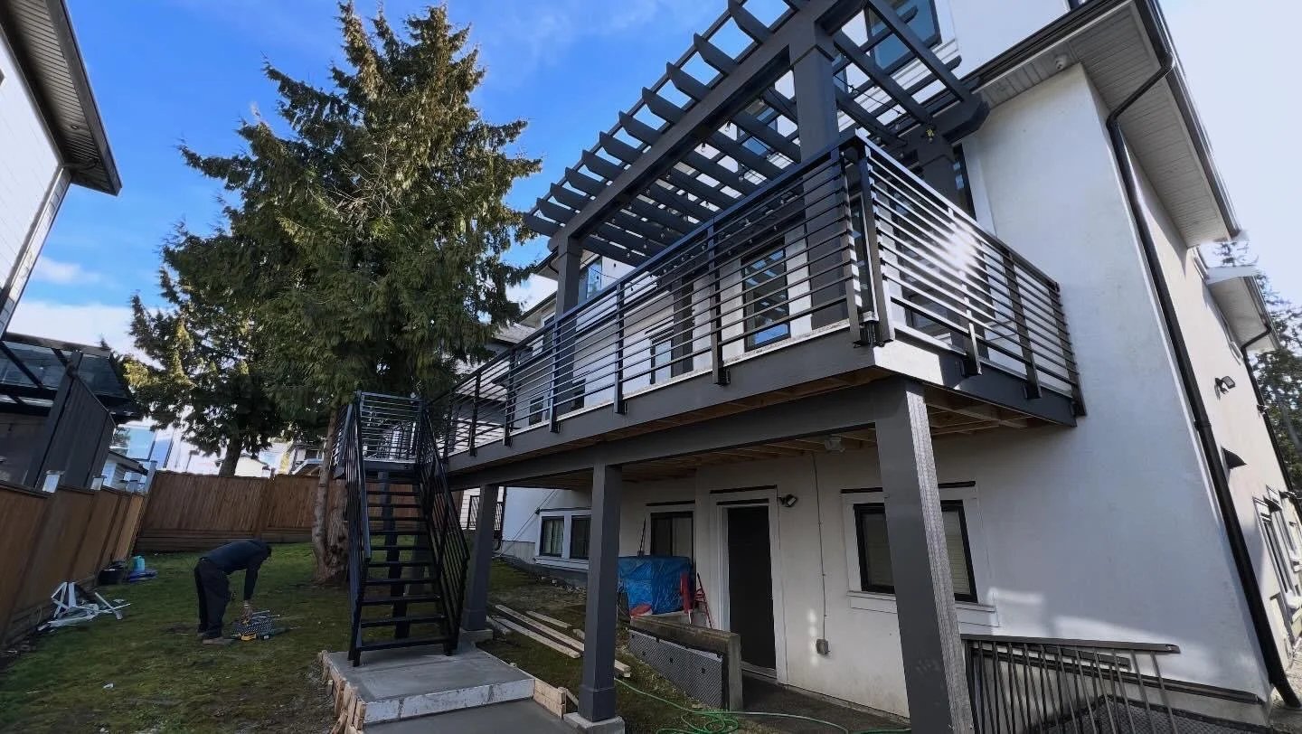 Construction worker installing a new black metal staircase leading to the second-floor balcony of a modern white house. The house has stacked black railings on the balcony and a large tree beside it, with tools and building materials on the grassy ya