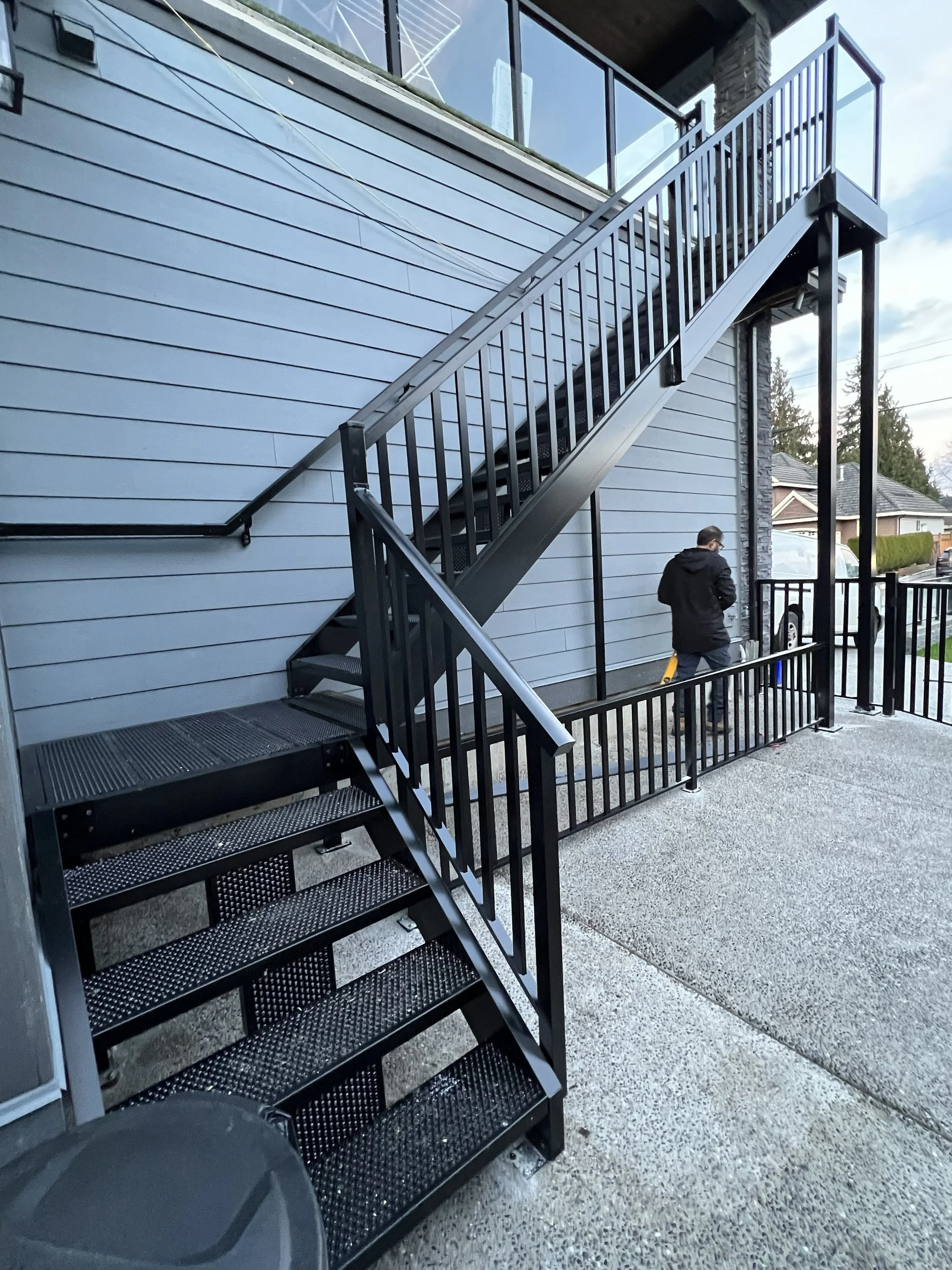 A metal outdoor staircase attached to the side of a house with blue siding and a stone accent wall. The staircase has black railings and steps, and a person in a black jacket is walking underneath it. The scene is set in a residential neighborhood wi