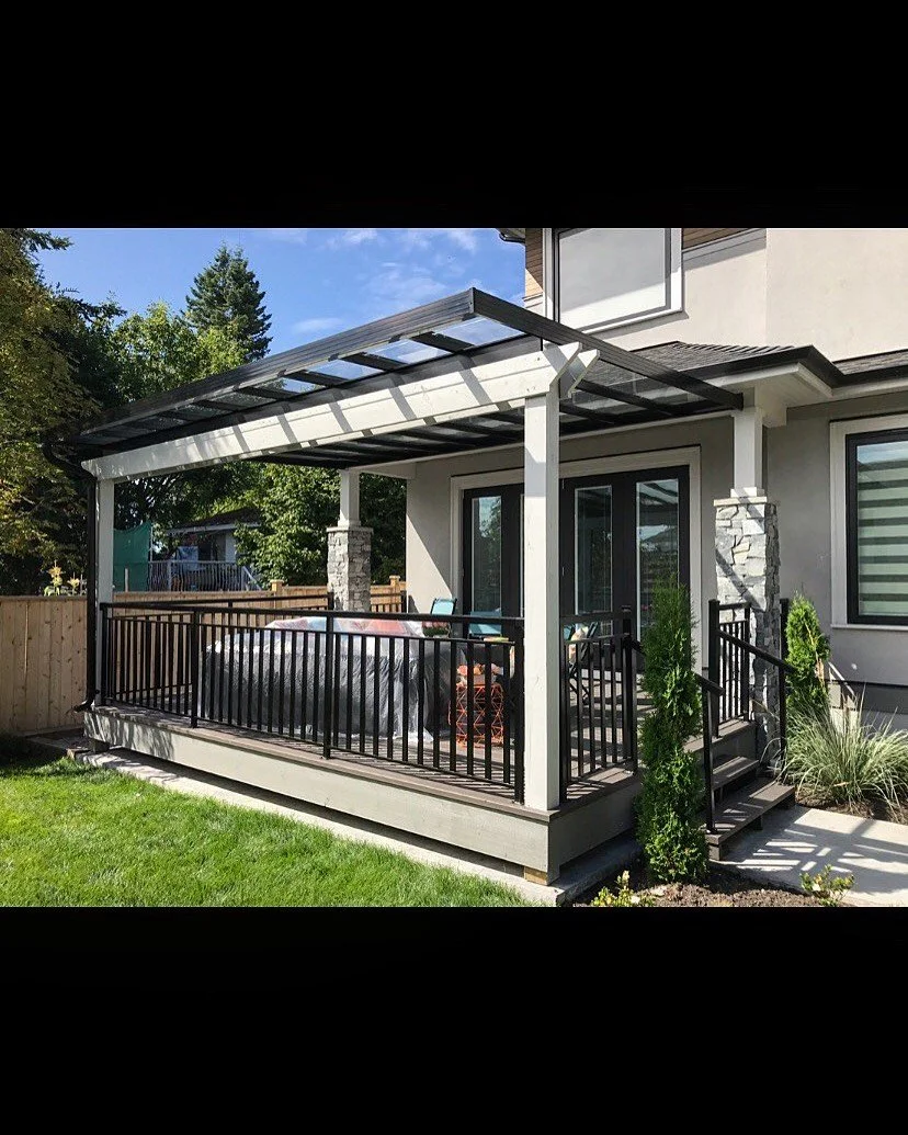Backyard patio with a wooden deck, black railing, and a transparent roof cover, connected to a house with sliding glass doors, surrounded by green lawn and plants.