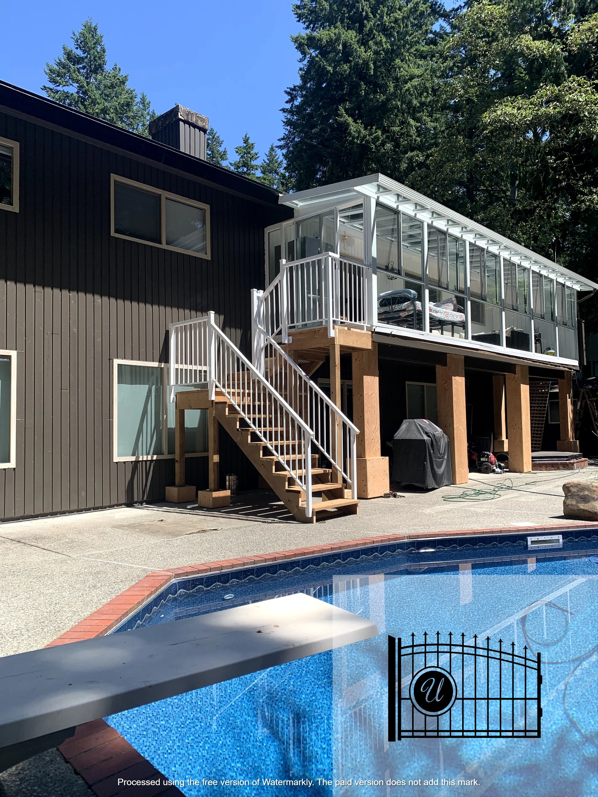 A house with a new screened porch built on the upper level, with outdoor stairs leading to it. There is a swimming pool in the foreground, surrounded by a concrete patio, and tall trees in the background.