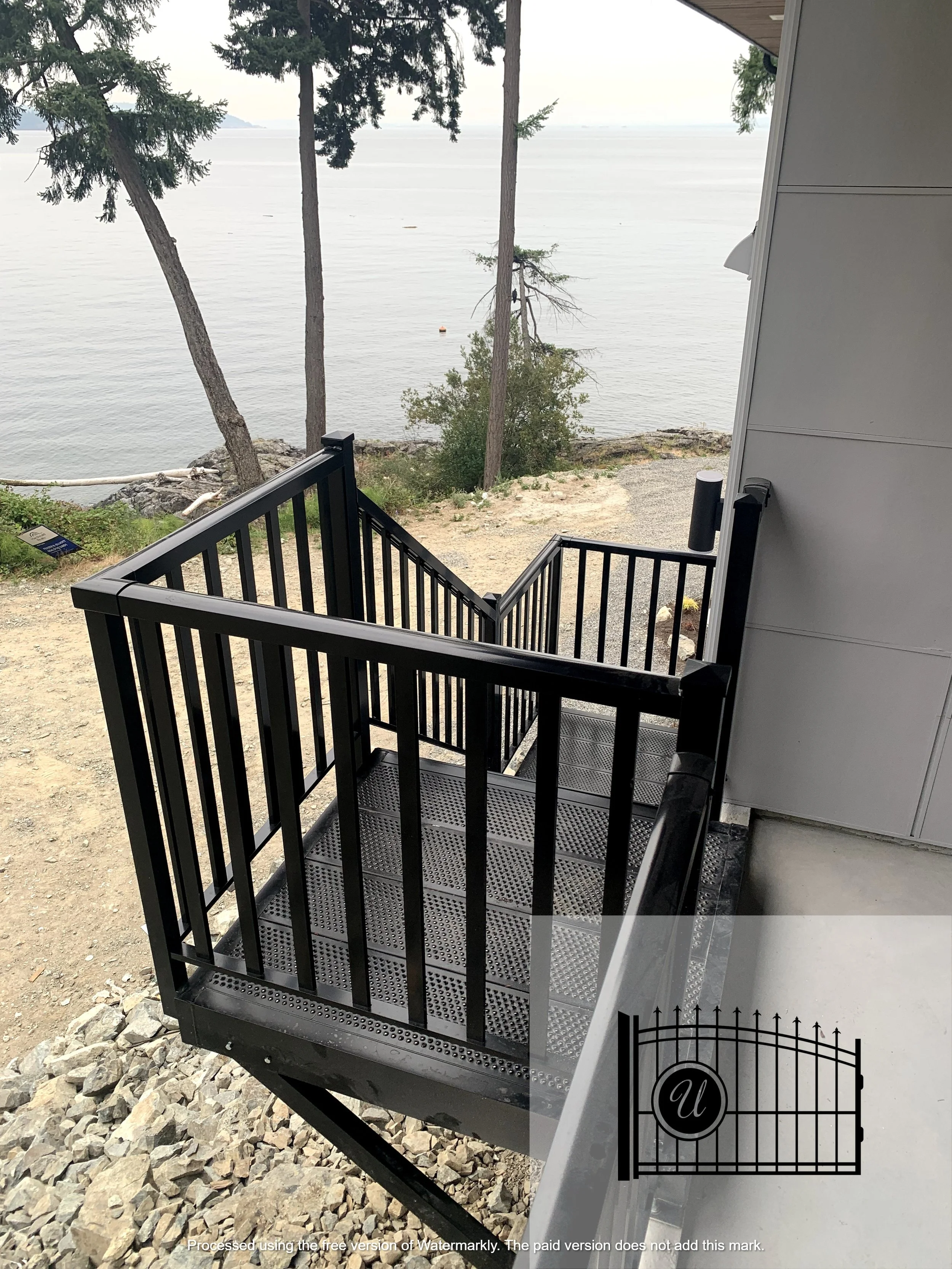 Black metal staircase with railings leading from a building to a rocky outdoor area near a body of water. In the background, there are trees and a calm water scene with distant landforms.