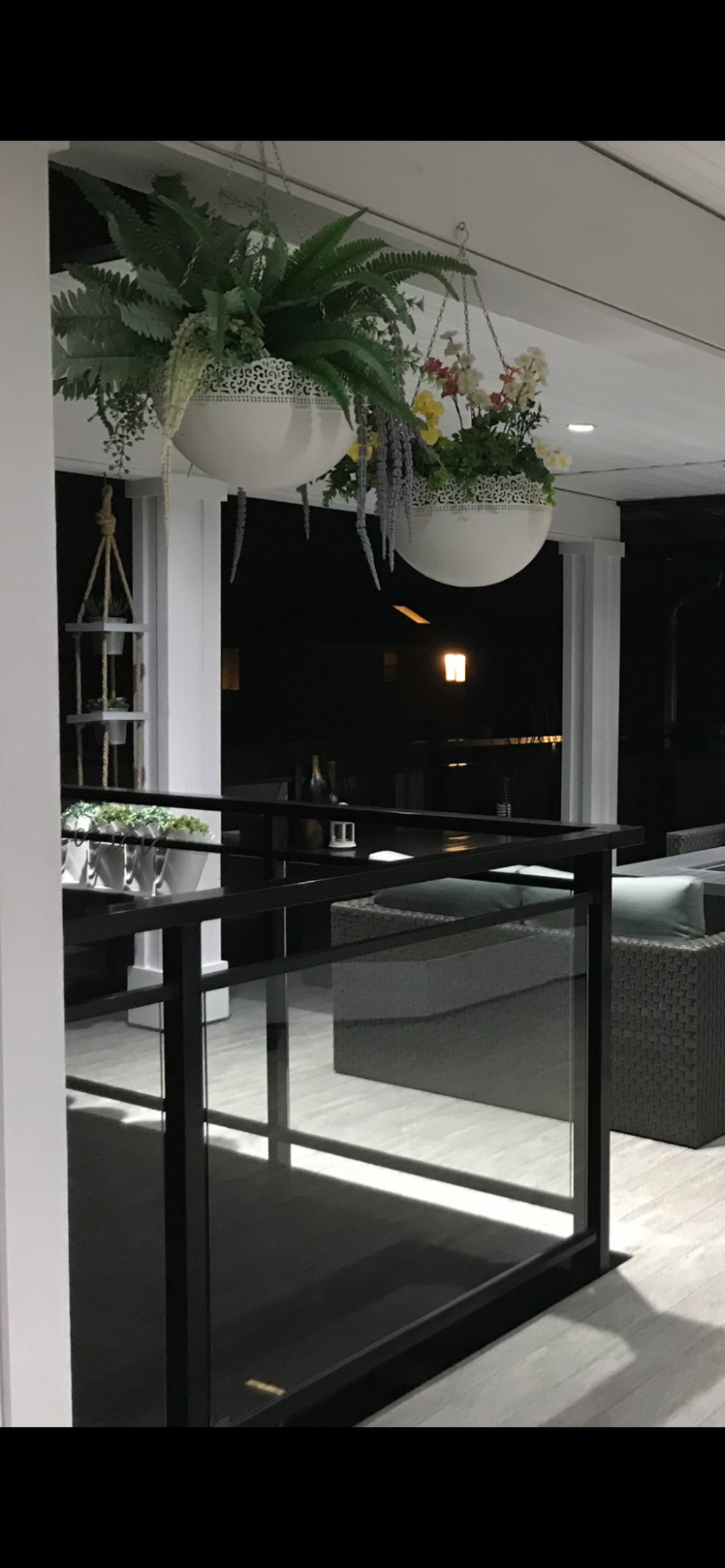 Nighttime outdoor patio scene with hanging planters containing green ferns and flowering plants, wicker chairs, and decorative items on tables.