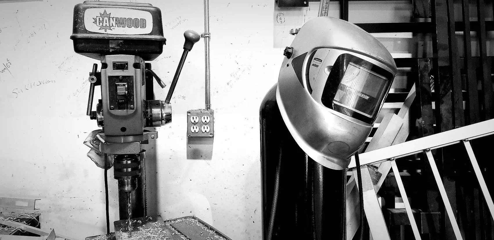 Black and white photo of a Coronet drill press, a welding helmet, and a power outlet in a workshop or garage setting.