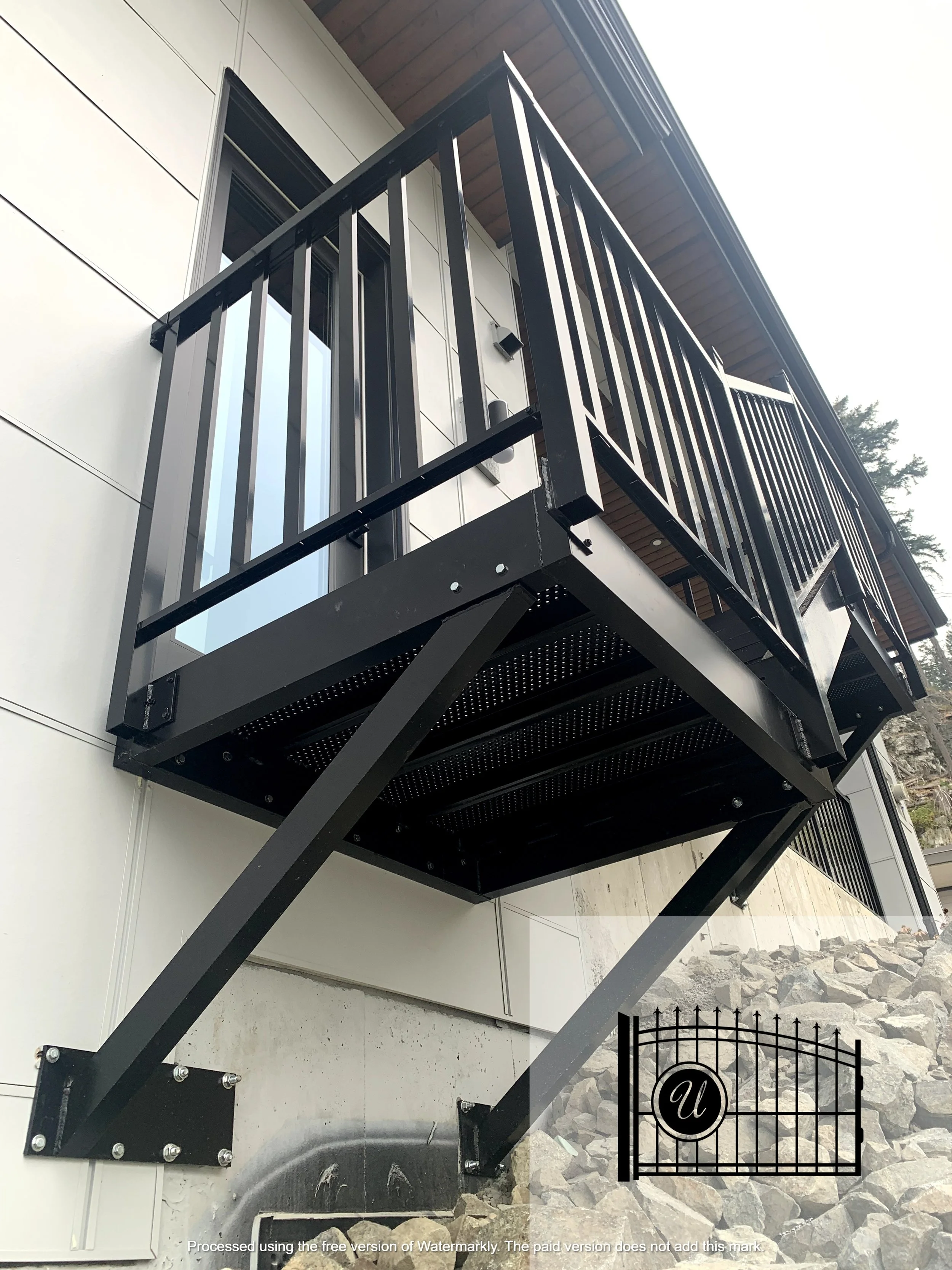 Black metal balcony railing attached to the exterior wall of a building, with a small gate at the bottom, against a background of trees and rocky ground.