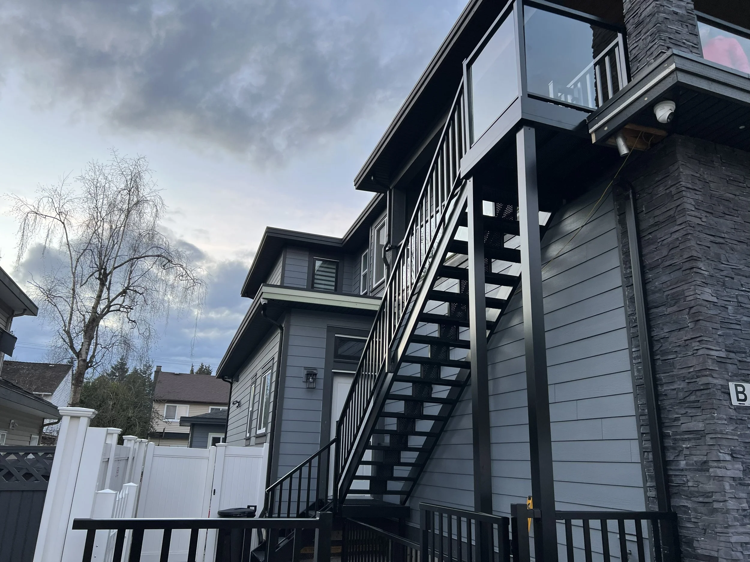 A two-story house with gray siding and a stone accent wall, featuring a black metal exterior staircase leading to the upper level, surrounded by a white privacy fence with a black gate, and a leafless tree against a cloudy sky.