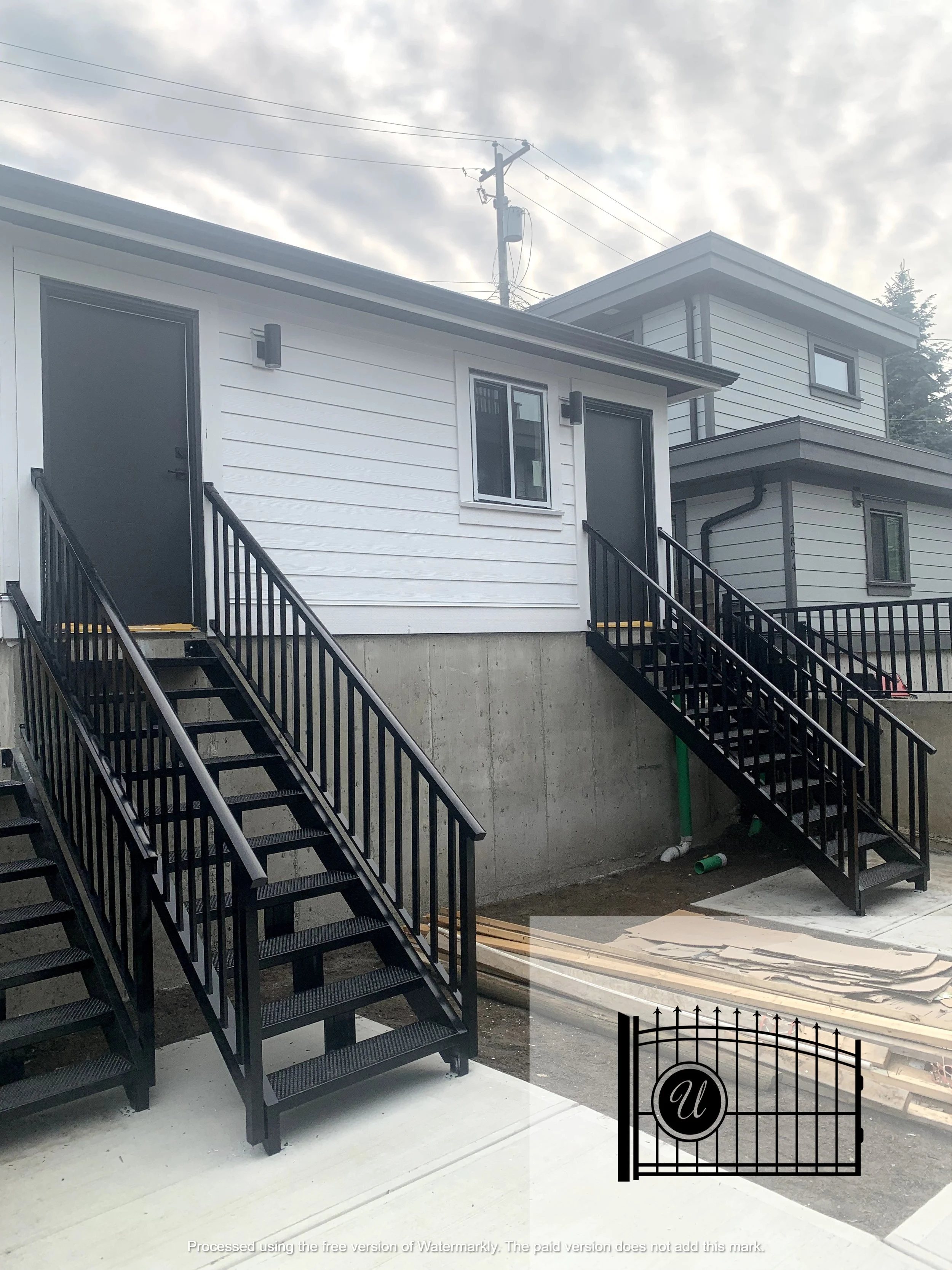 Exterior of modern two-story houses with black metal staircases and black handrails, a utility pole with wires, and a cloudy sky.