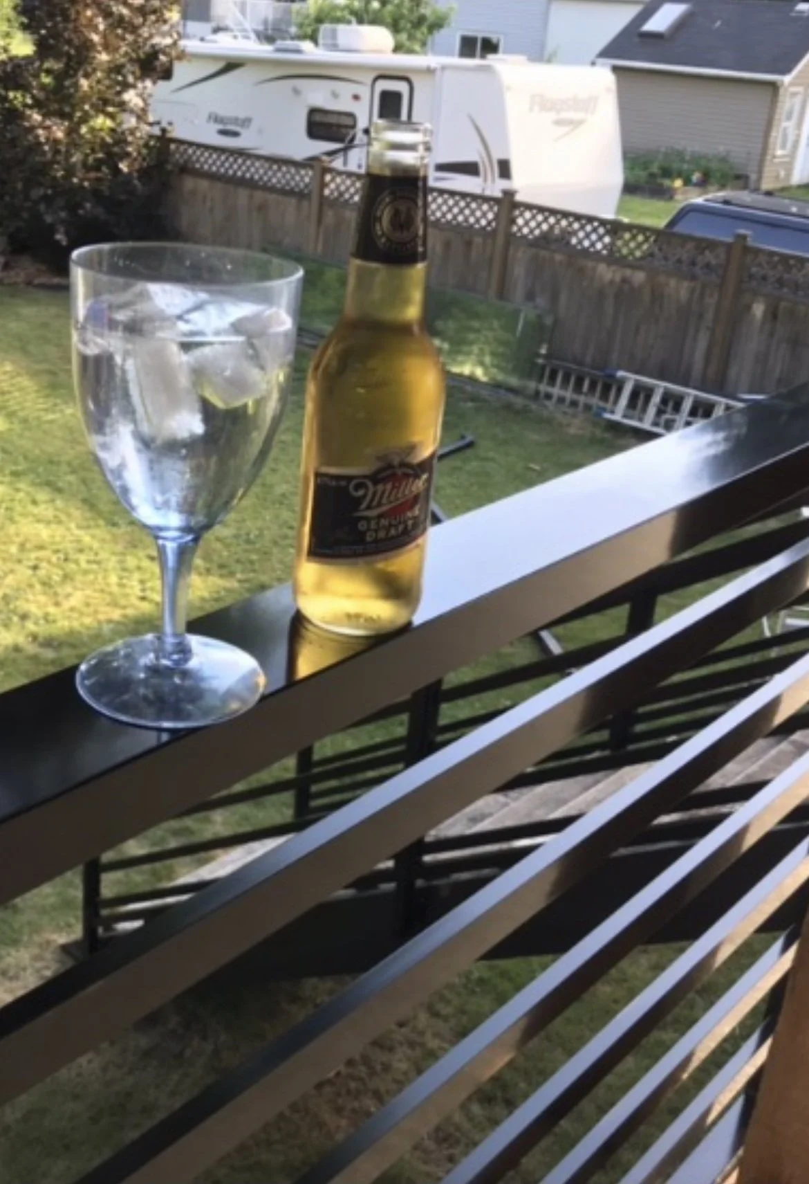 A glass of water with ice and a bottle of Miller beer on a balcony railing, overlooking a backyard with a garden, wooden fence, a ladder, and a RV in the background.