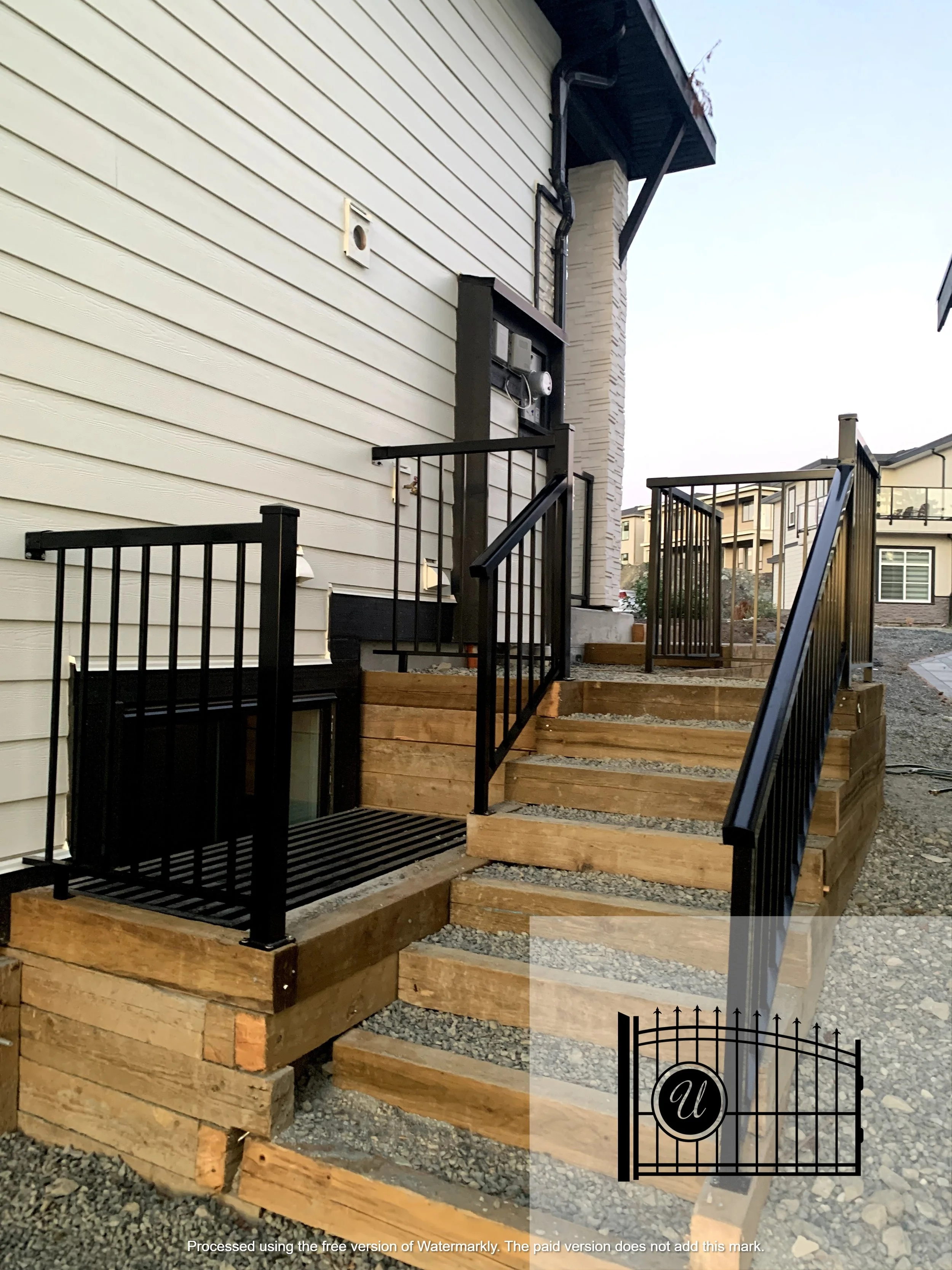 A newly constructed outdoor staircase with black metal railings leads up to a house with beige horizontal siding. The stairs rest on wooden supports and the area around the stairs is gravel. There is a black gate at the bottom of the stairs.