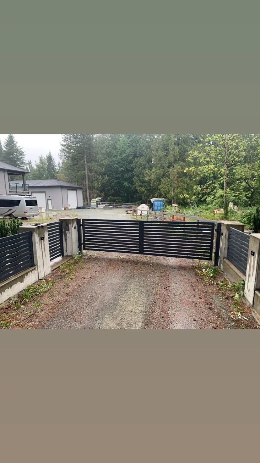 Driveway with black metal gate and concrete pillars, leading to a rural property with trees and buildings in the background.