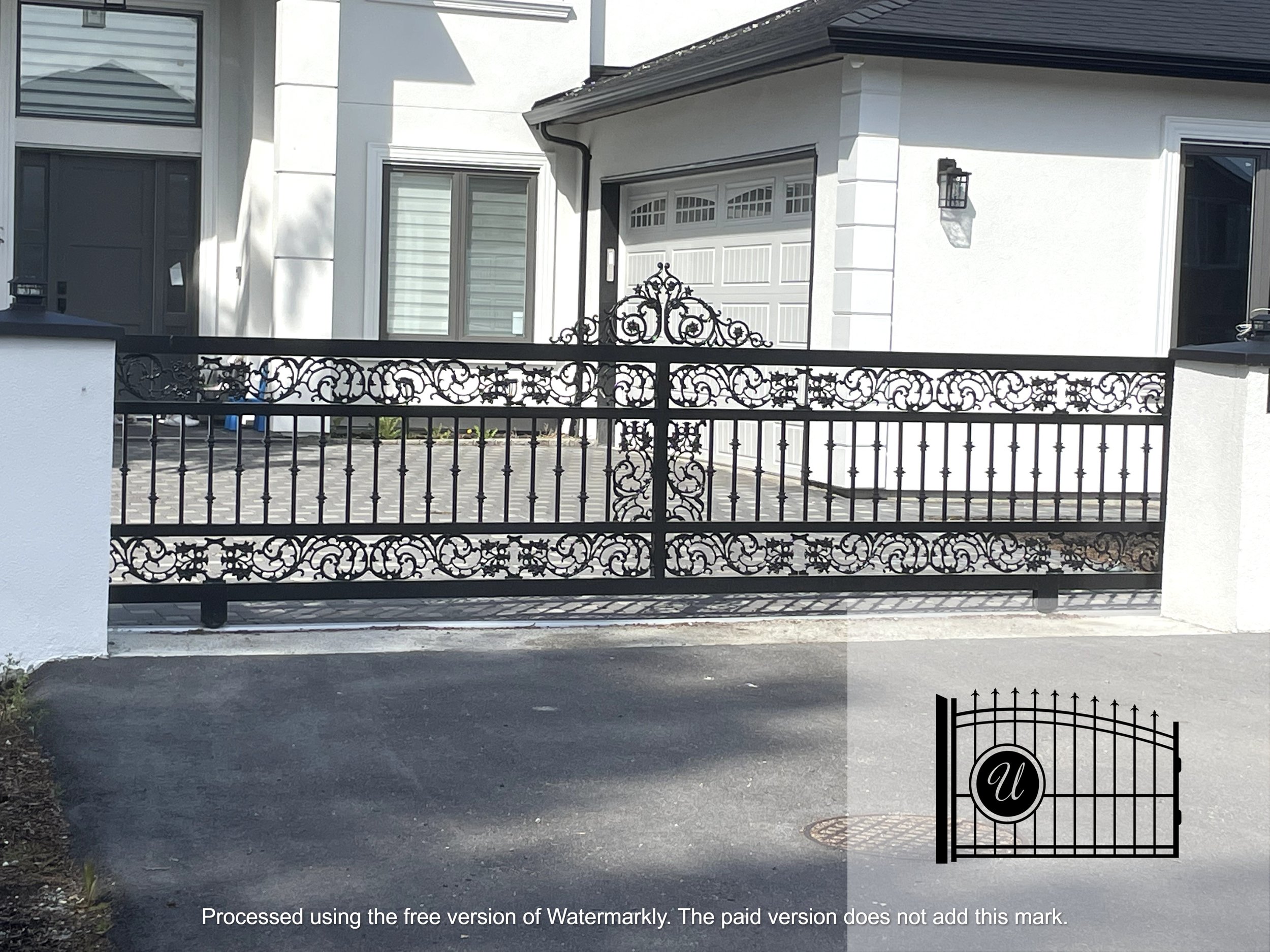 White house with black wrought iron gate in front, with a driveway leading up to a white garage door with windows. There is a lit outdoor wall lantern near the garage and a larger window to the right.