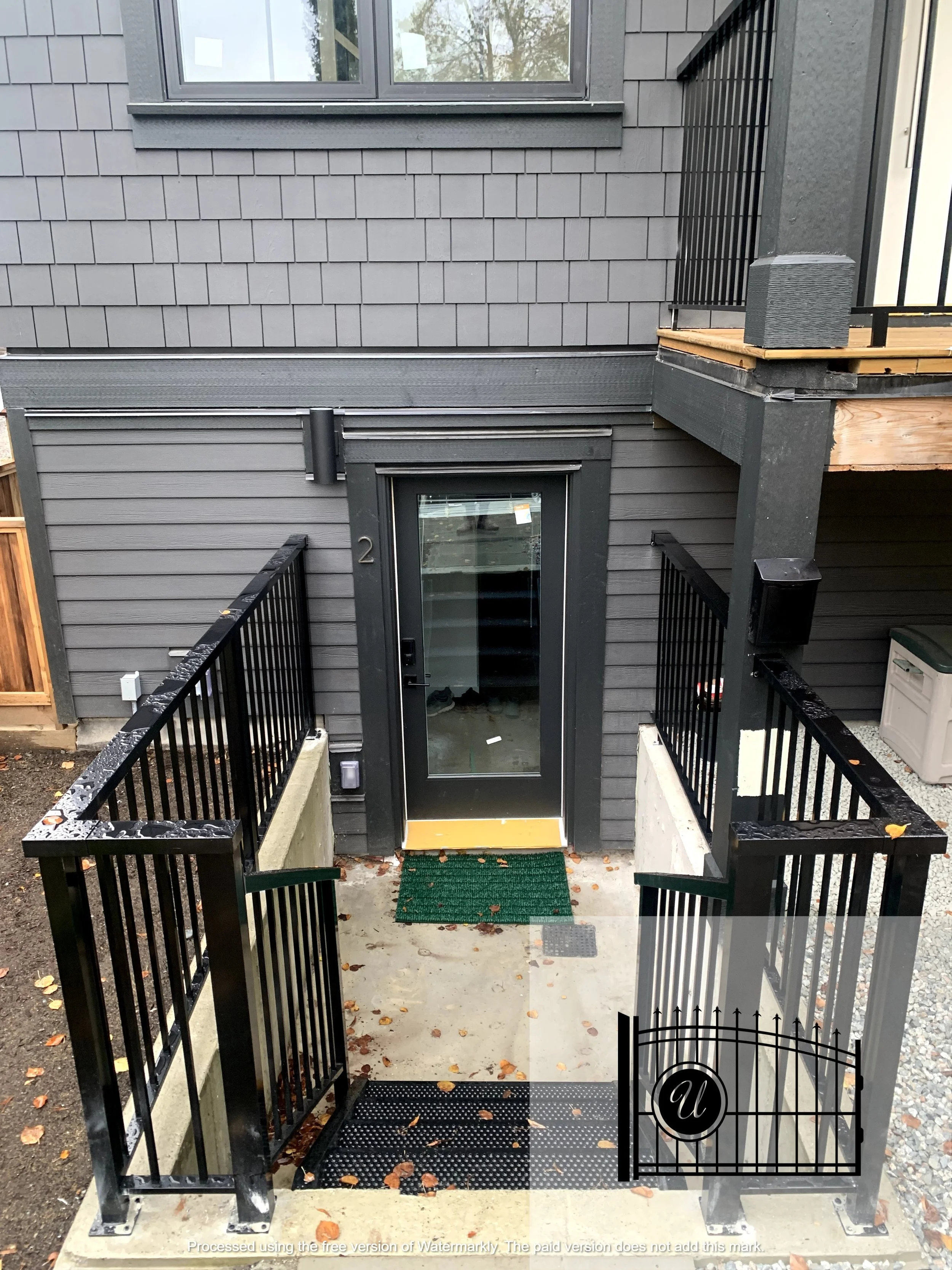 Black metal gate with 'U' shape and decorative elements in front of a gray house entrance, with a glass door, black railings, a green mat, and fallen autumn leaves.