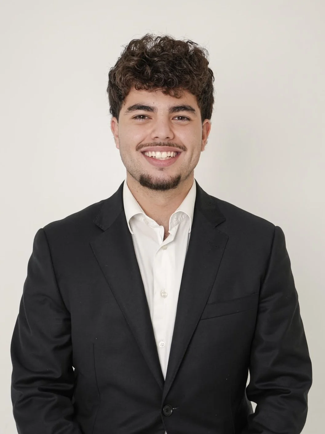 A young man with curly brown hair and a goatee, wearing a black suit jacket and white dress shirt, smiling against a plain off-white background.