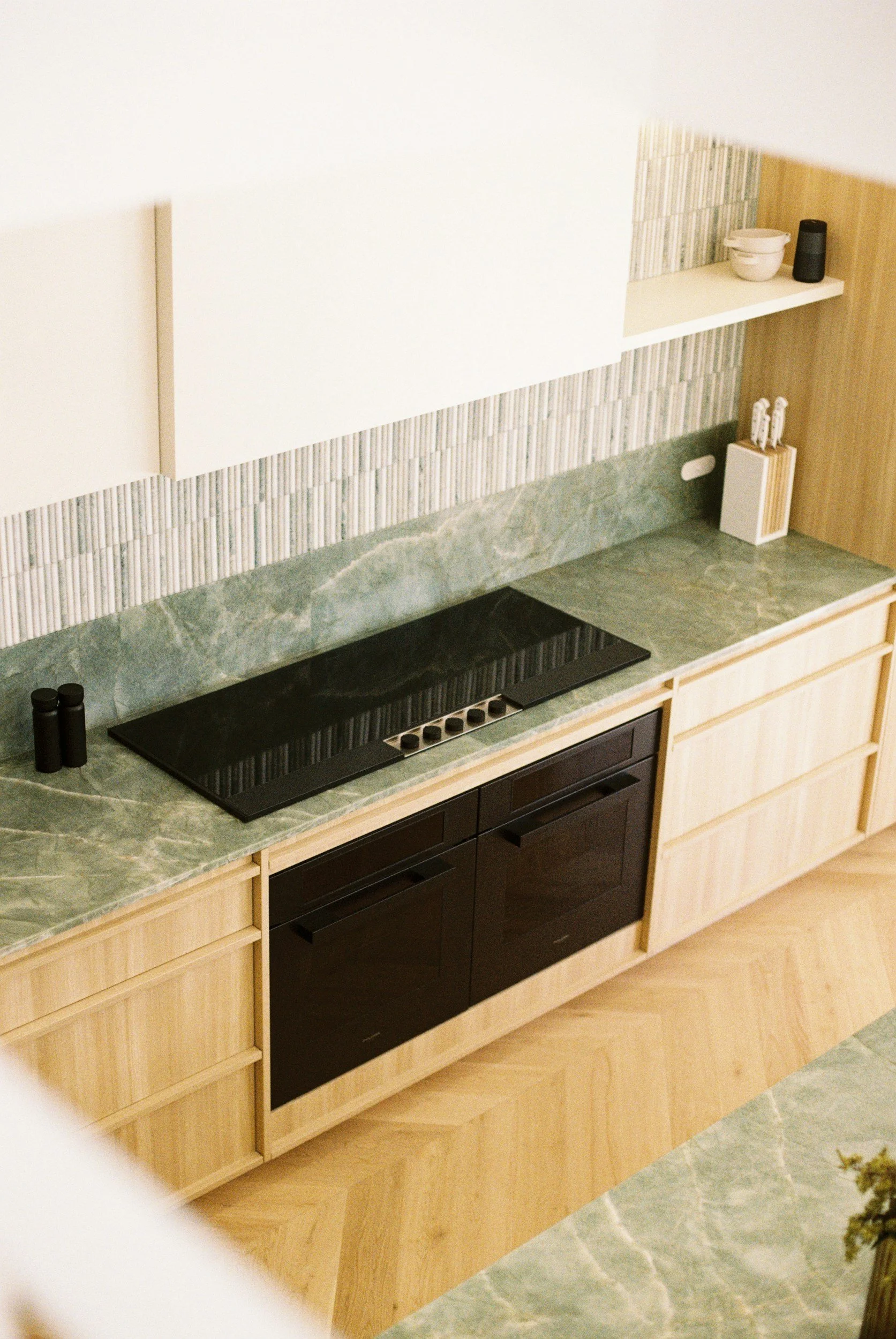 Close-up photo of a modern kitchen with wooden cabinets, a black stove, and a greenish marble countertop, with a partial view of a backsplash.
