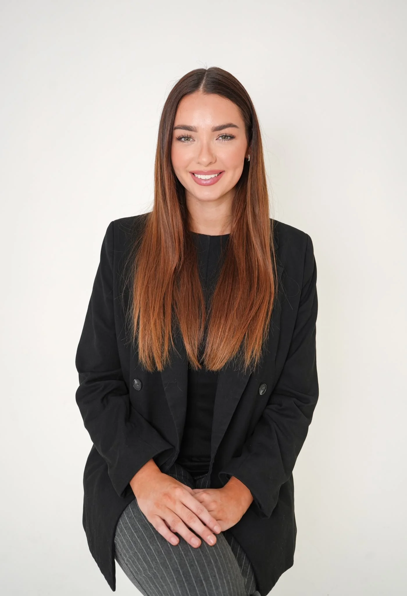A woman with long reddish-brown hair, wearing a black blazer and pinstripe pants, sitting against a plain white background, smiling with her hands crossed on her lap.
