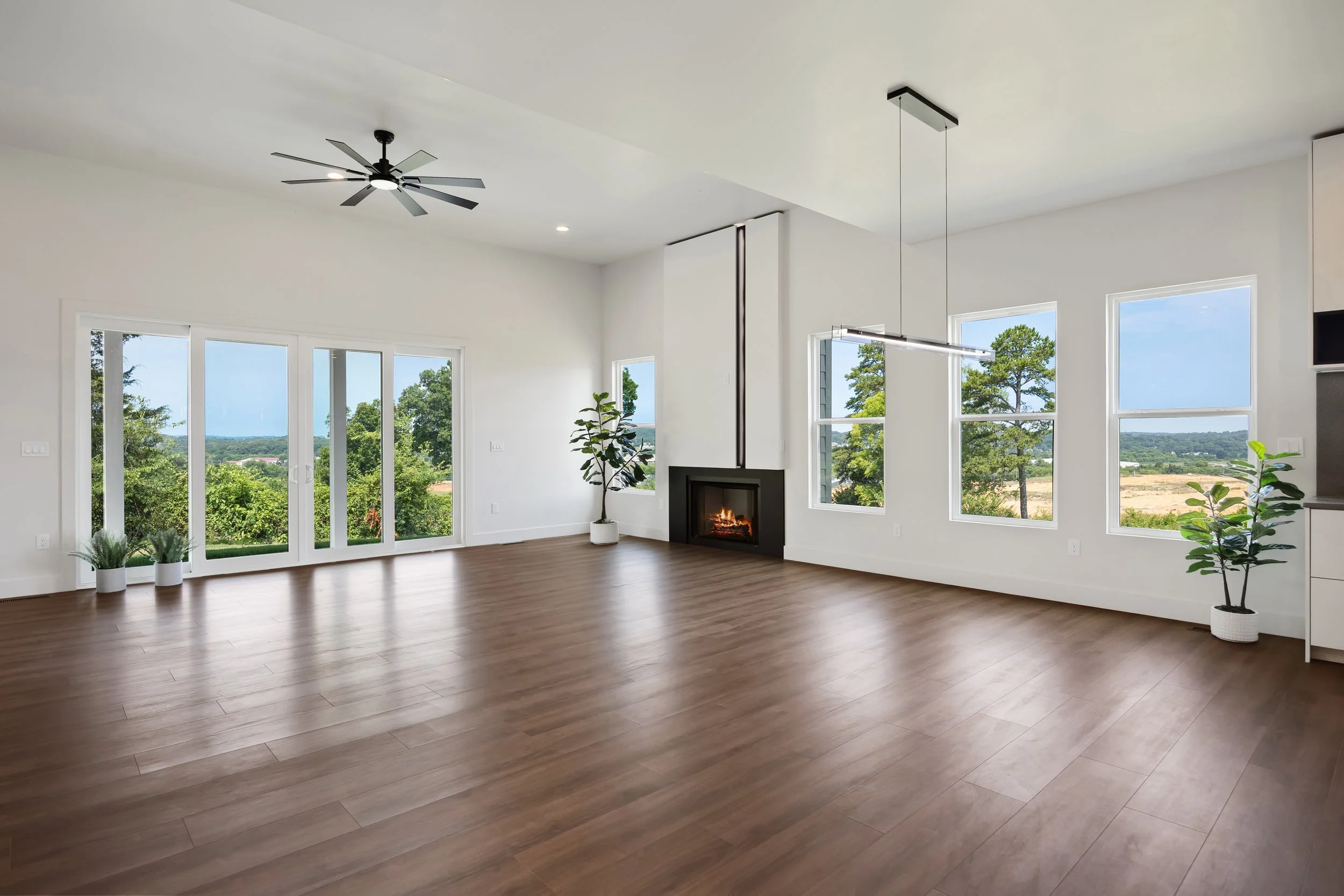 A spacious, modern living room with white walls, wood flooring, large windows showing greenery outside, a ceiling fan, a fireplace, and potted plants.