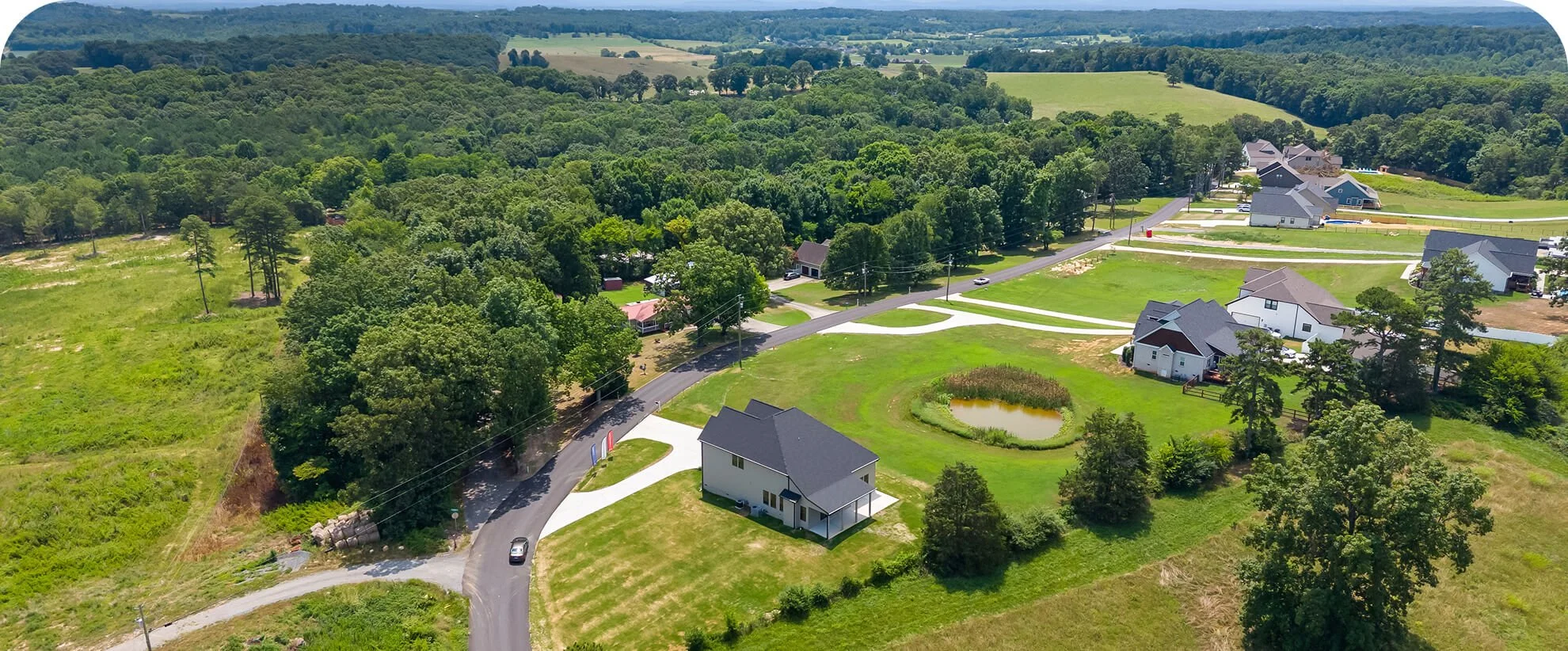 Aerial view of a suburban neighborhood with houses, greenery, and a small pond, surrounded by trees and hills in the distance.