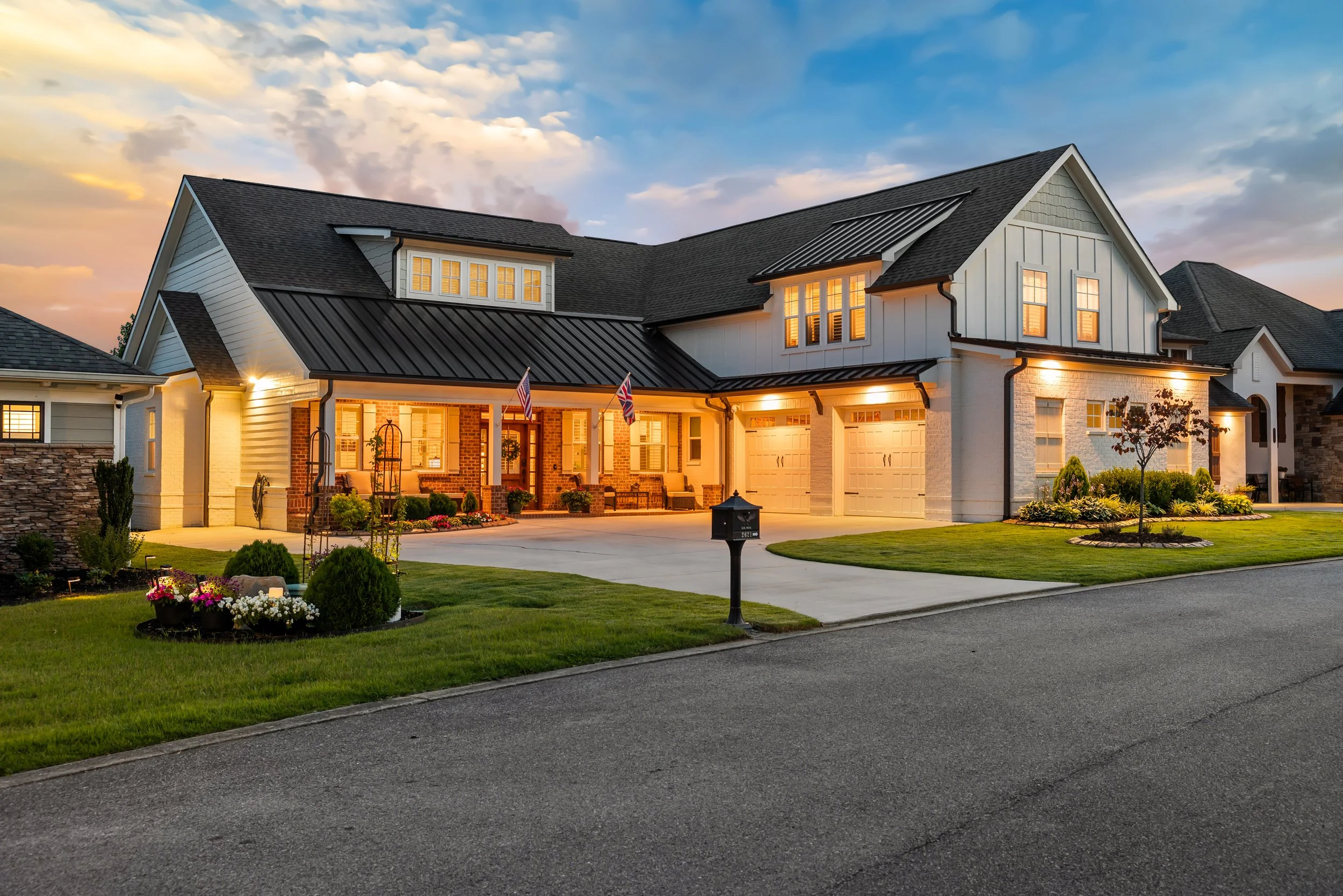 A large suburban house during sunset with illuminated windows and a well-kept lawn. The house has a mix of modern and traditional design with a prominent front porch and multiple gables, flags near the entrance, and a driveway leading to a garage.