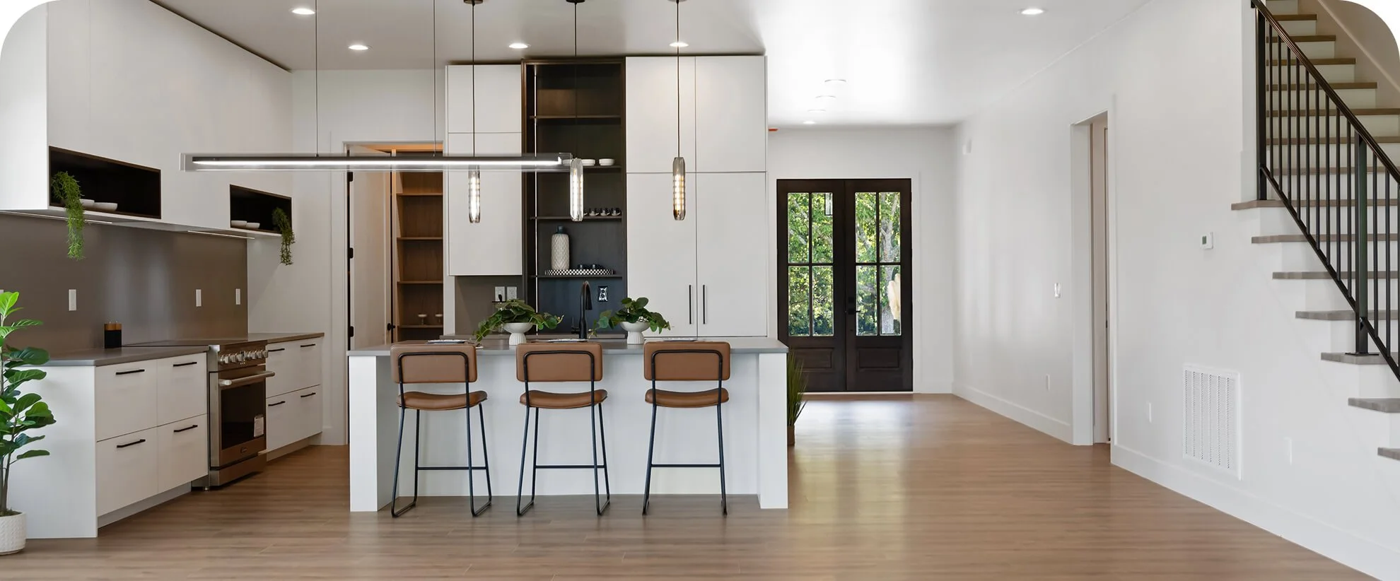 Modern kitchen with white cabinets, black accents, and a center island with four brown bar stools. There are black double doors at the entrance, and a staircase with black railing on the right. Natural light illuminates the space, and there are green plants for decoration.