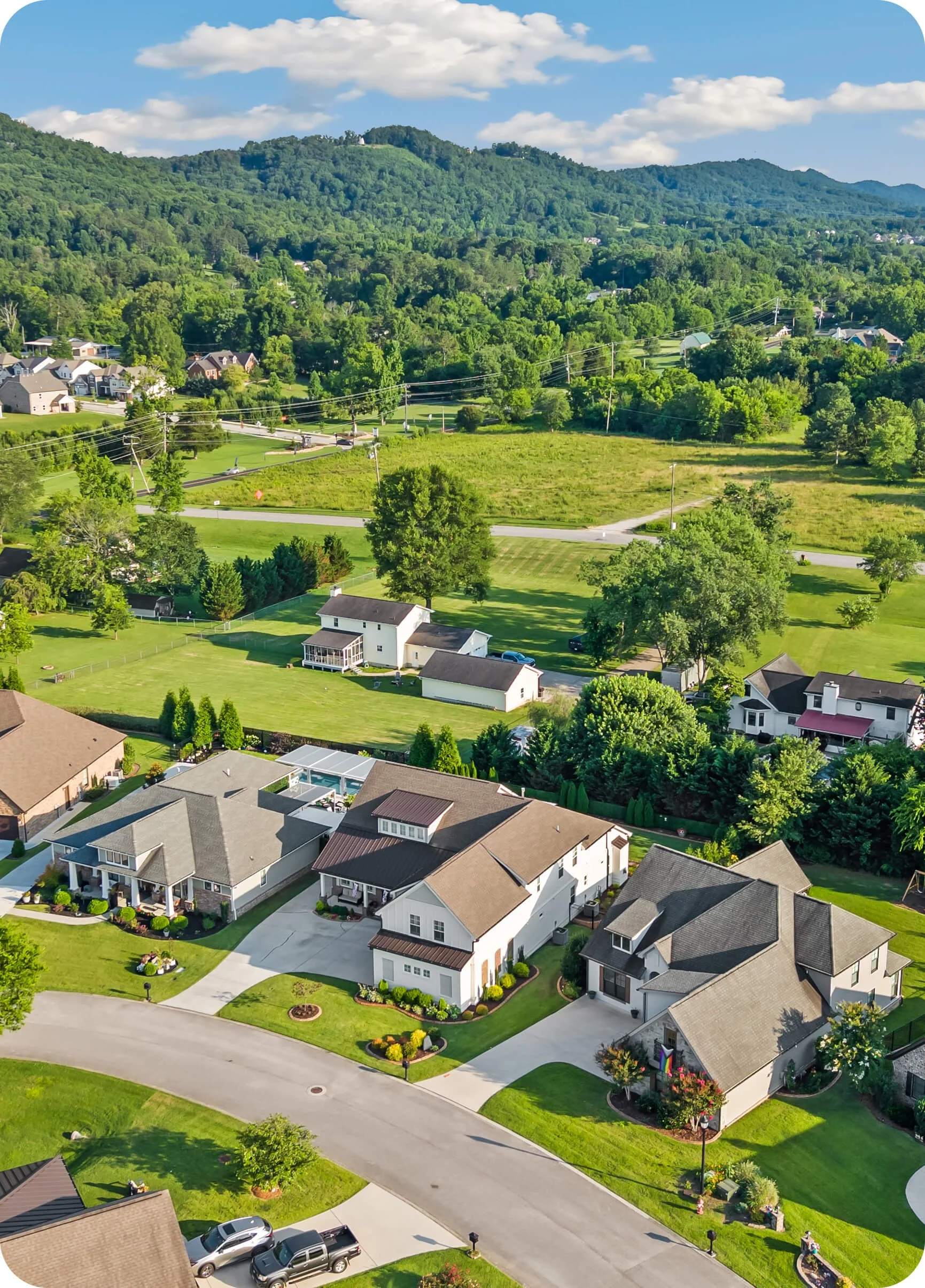 Aerial view of a suburban neighborhood with houses, green lawns, trees, and a backdrop of rolling hills and a blue sky with some clouds.