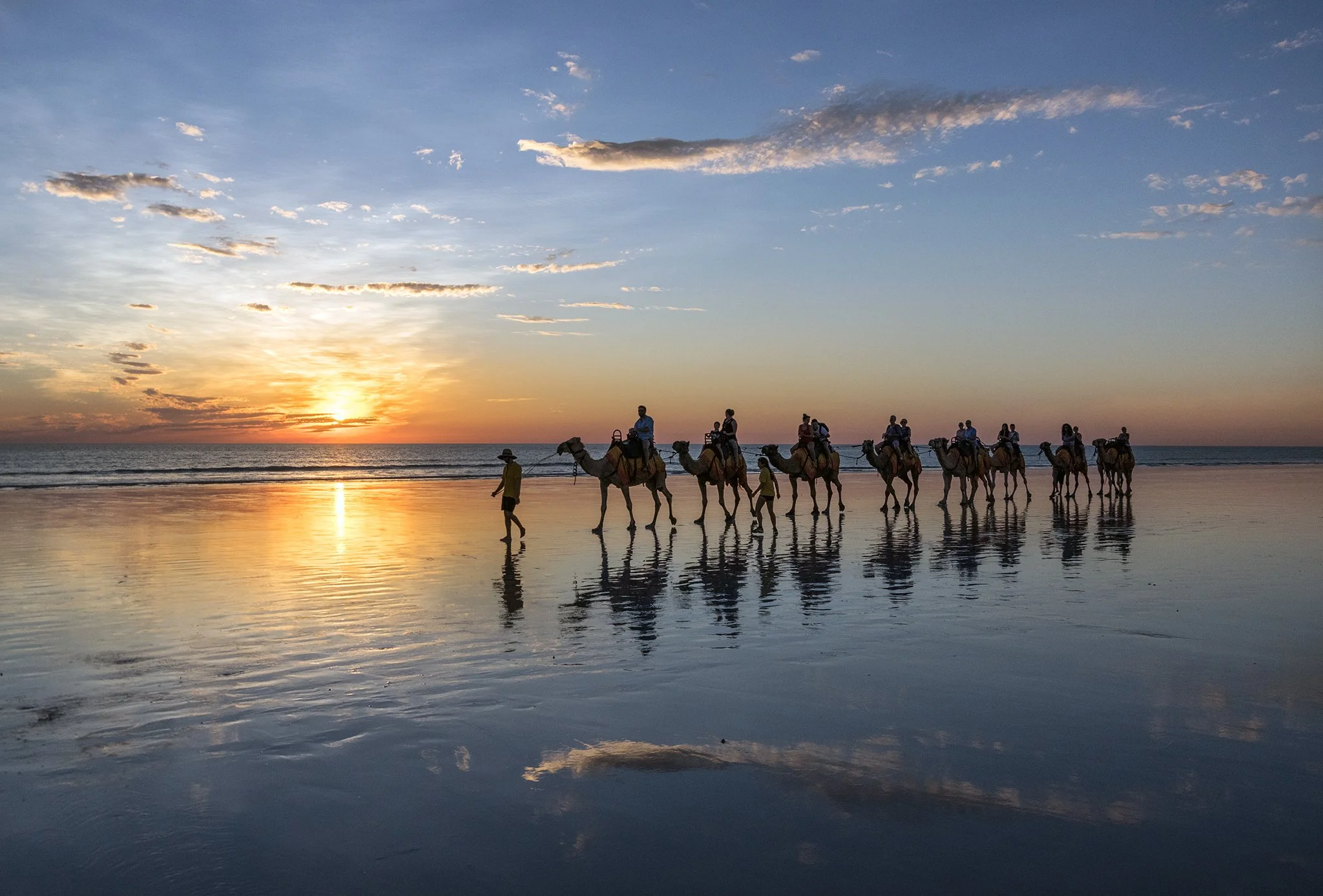 A group of people riding camels along the beach at sunset, with a reflection of the silhouettes in the wet sand.