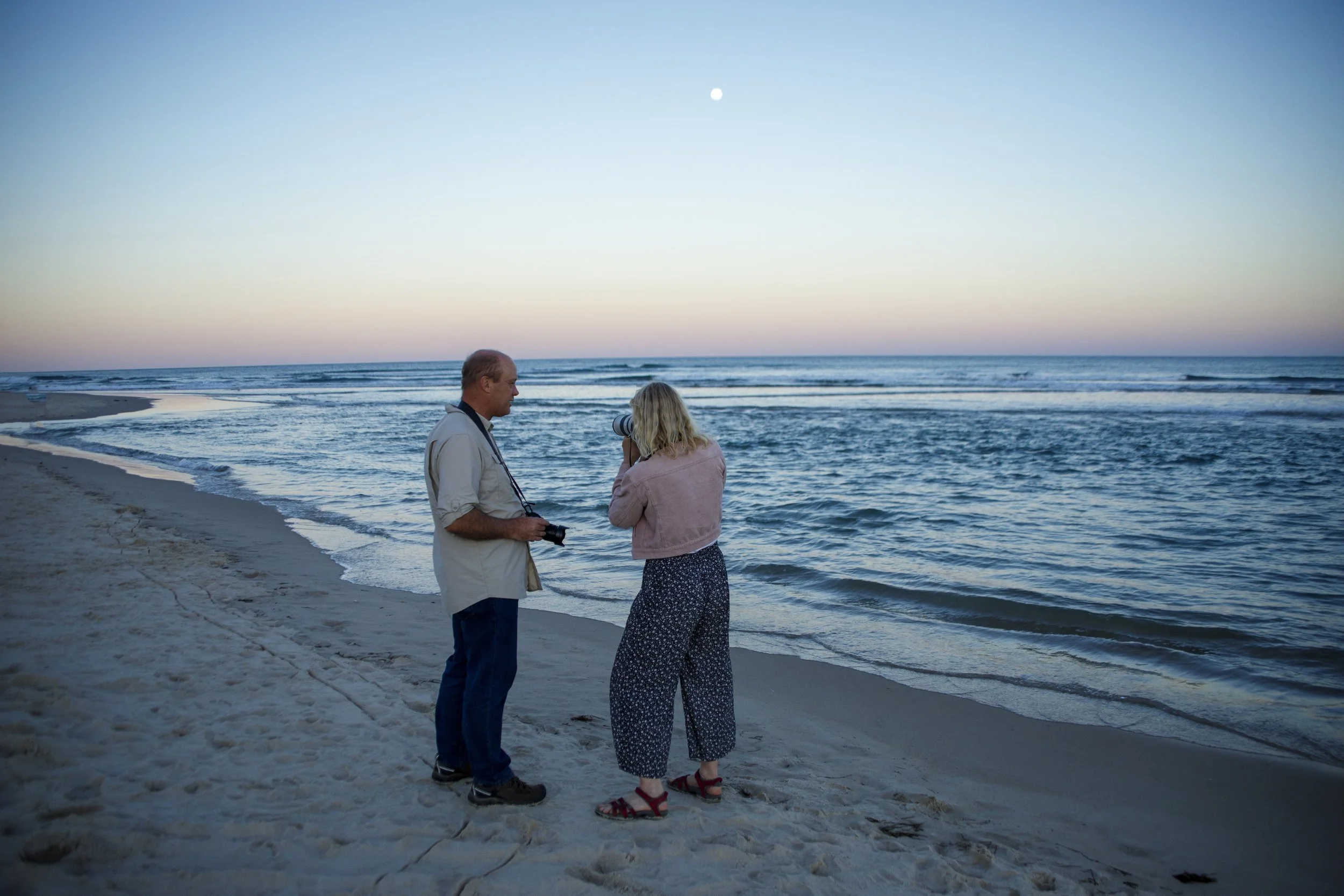 A man and a woman stand on the beach near the water. The man is holding a camera and the woman is looking through a telescope, both engaging in photography during sunset or dusk by the ocean.