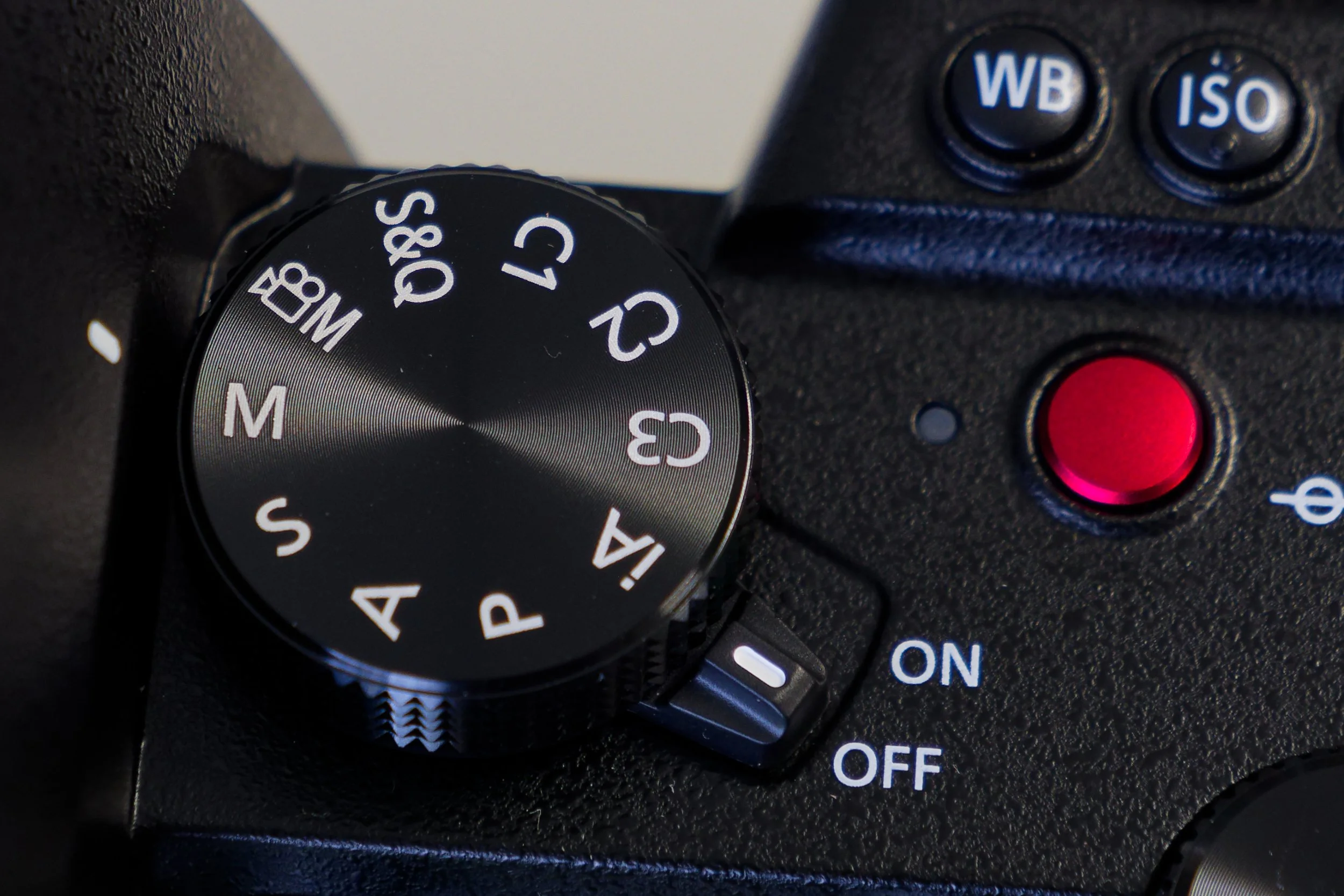 A close-up photograph of a camera's mode dial, featuring settings from 'A,' 'A+', 'S,' 'C,' 'M,' and others, with additional buttons labeled 'WB' and 'ISO' visible in the background.