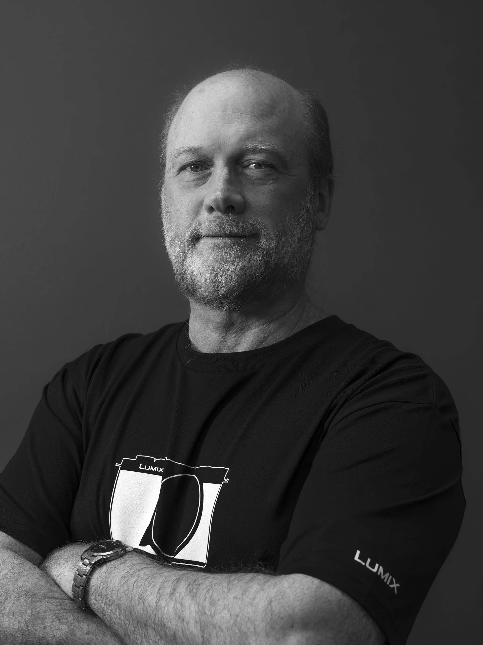 A black and white portrait of a middle-aged man with a beard and mustache, wearing a black T-shirt with a camera graphic and the word "LUMIX" on it, and a watch on his left wrist, against a solid gray background.
