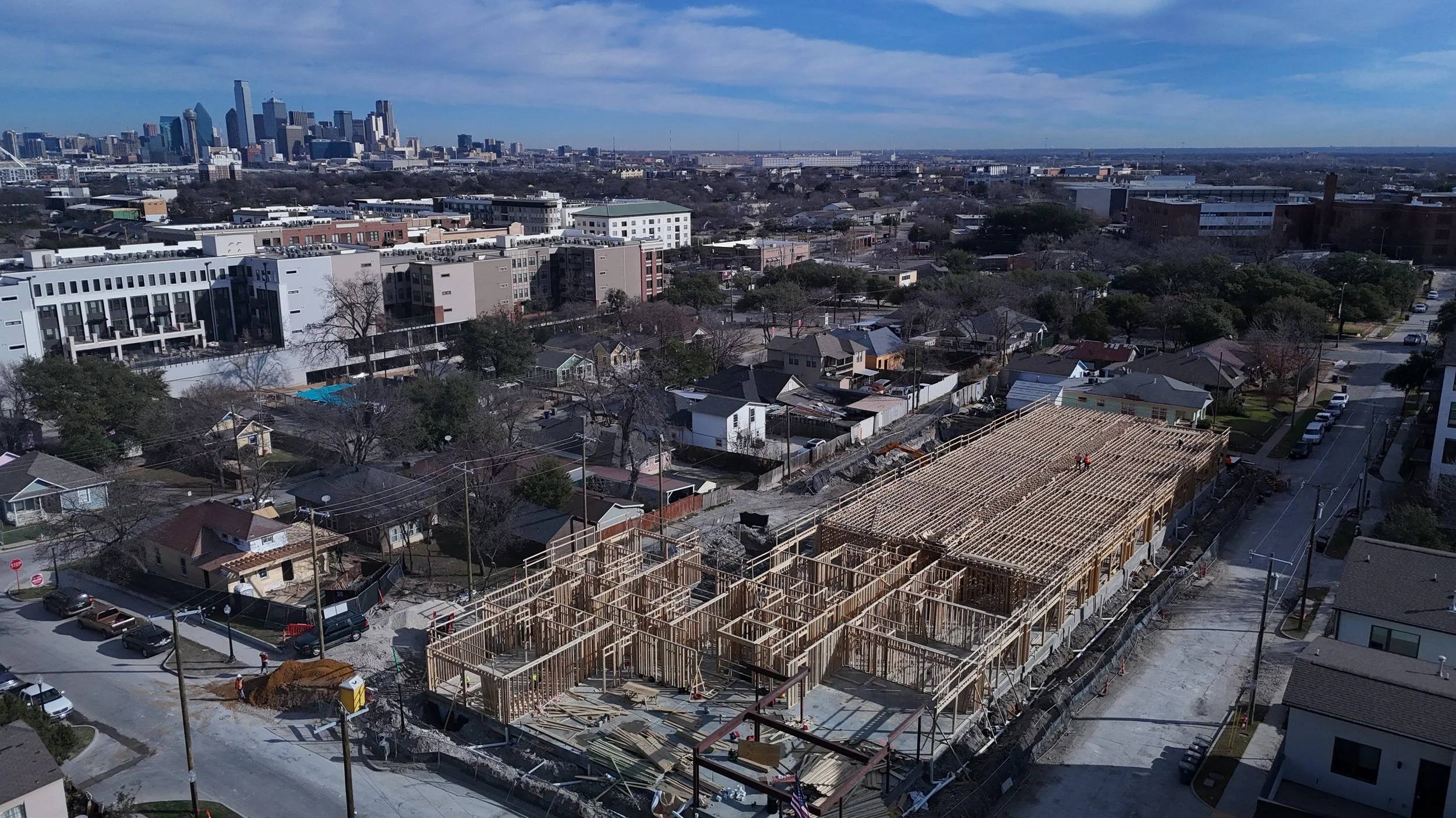 Aerial view of a wood-framed construction site with surrounding streets and a city skyline in the distance.