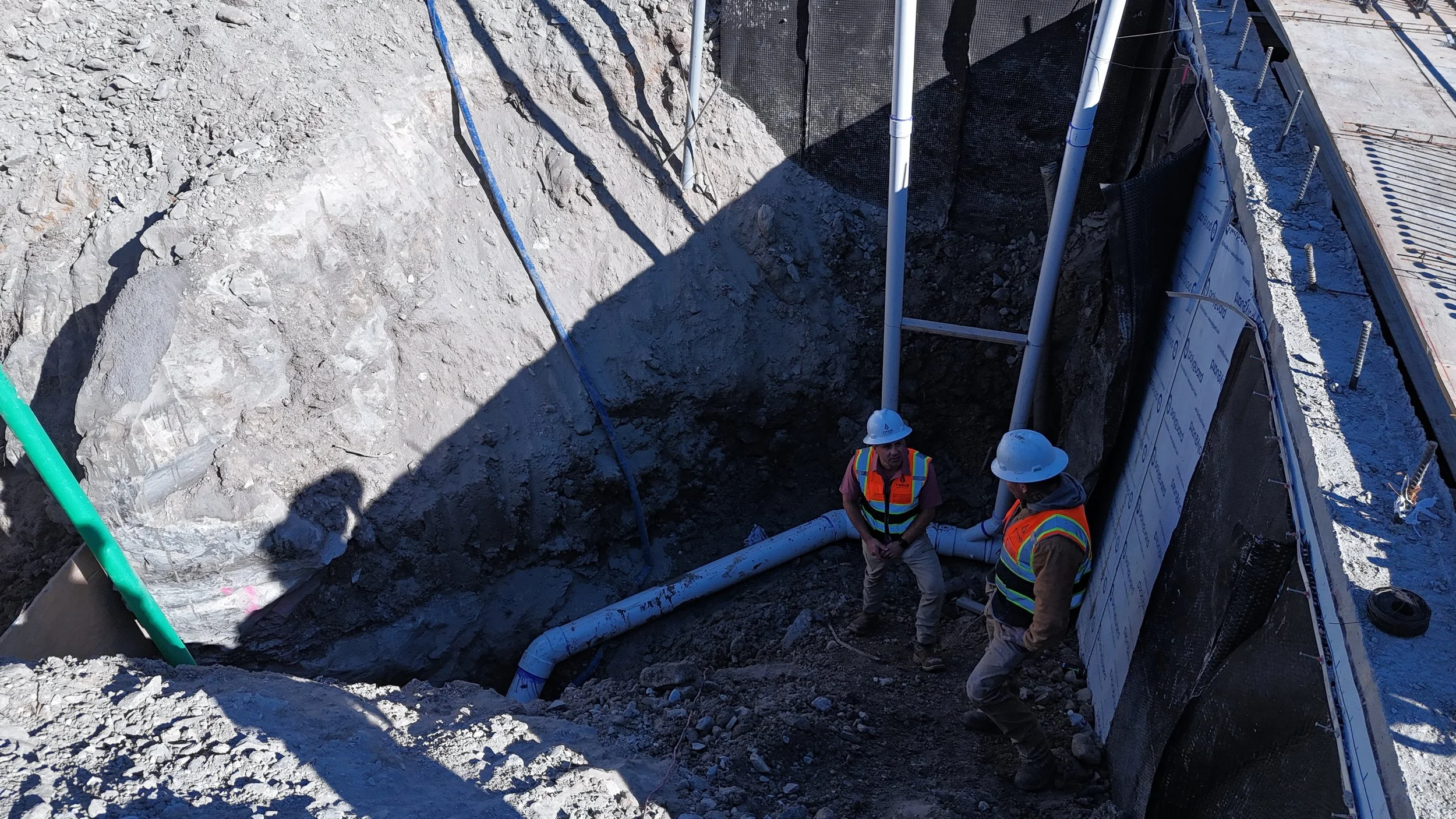 Two workers wearing hard hats beside vertical piping and a wall in an excavated construction area.