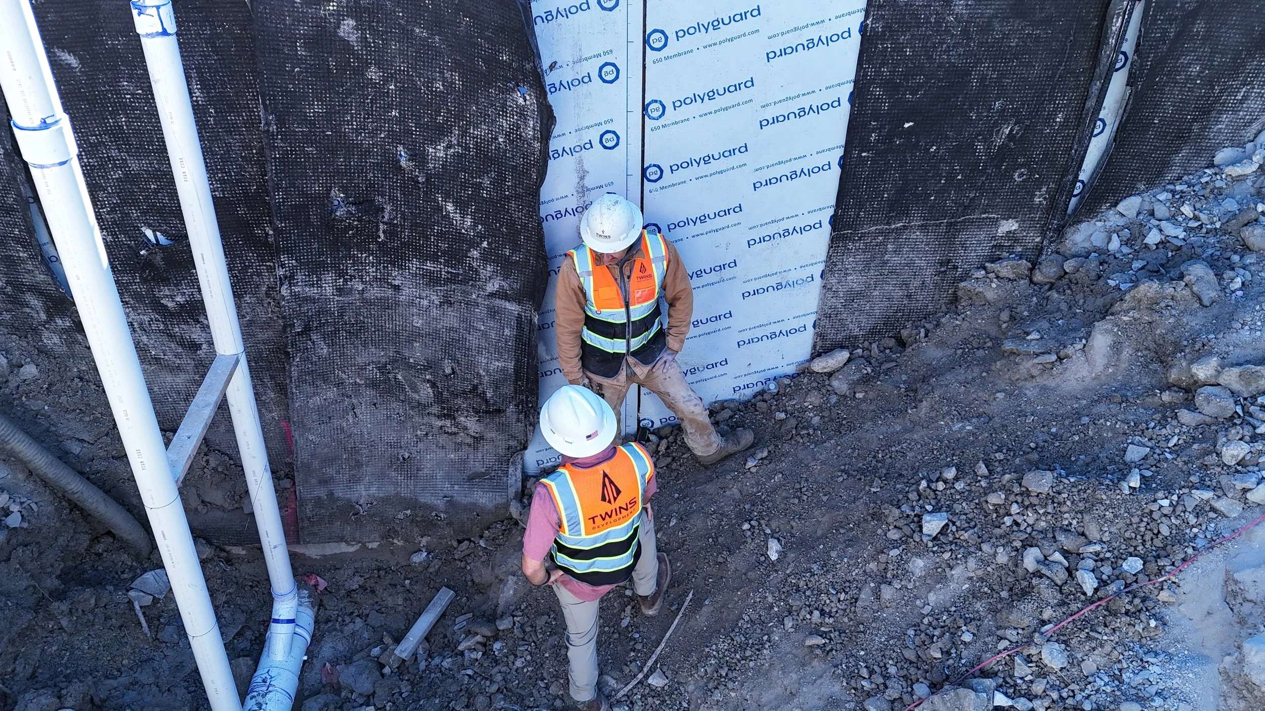 Two workers wearing hard hats and orange safety vests beside a covered rectangular column in an excavated area.